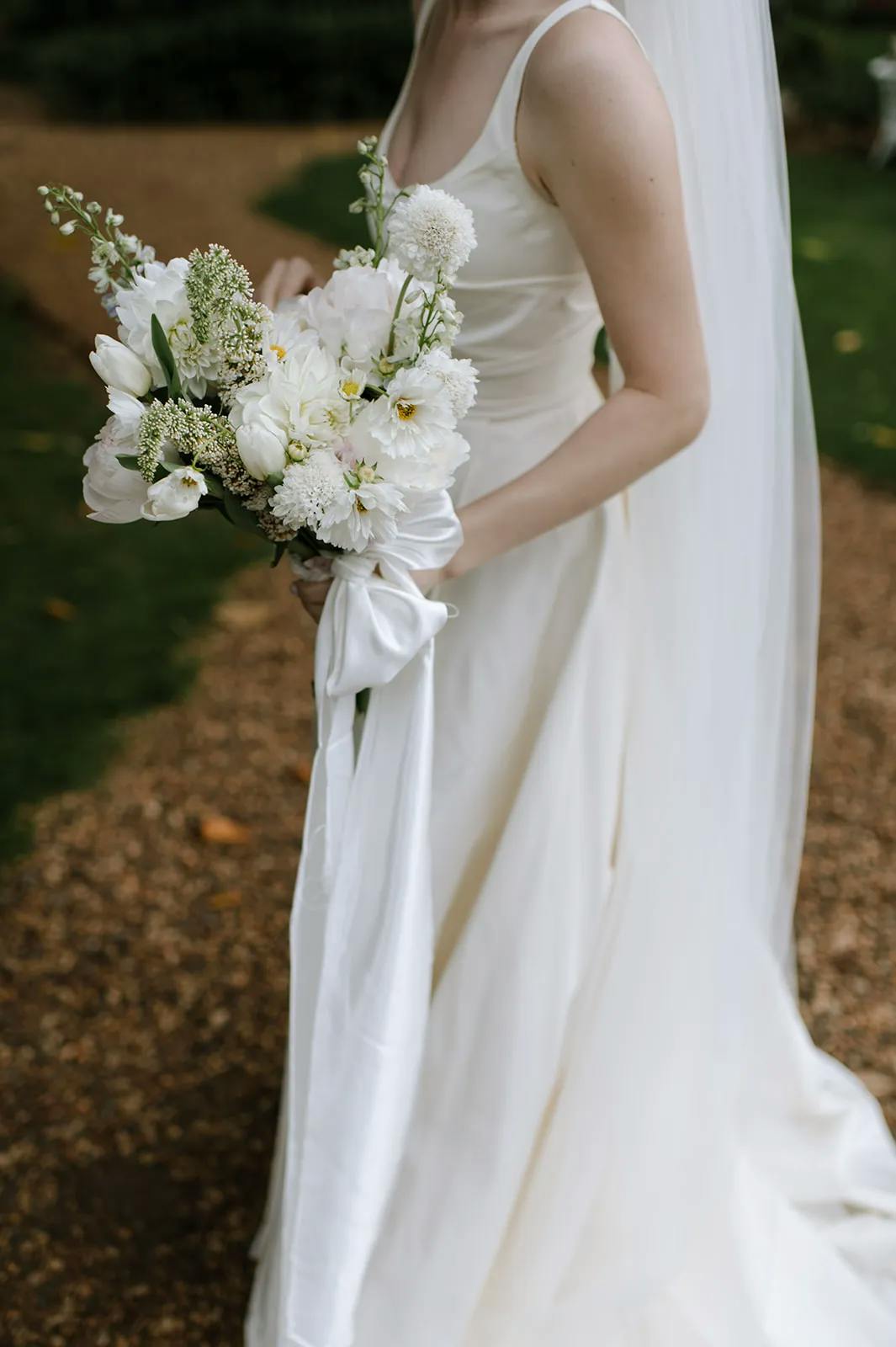 A bride in a sleeveless white wedding dress holds a bouquet of white flowers, including tulips, chrysanthemums, and greenery, wrapped with a white ribbon. The bride's long veil flows down her back, and the background features a garden path.