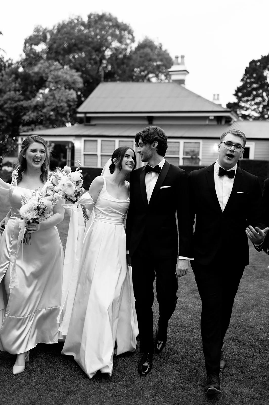 A black and white image shows four people walking together outdoors, dressed formally. The two women in white dresses carry bouquets, and the two men in black tuxedos are smiling and conversing. There's a building with a peaked roof in the background.