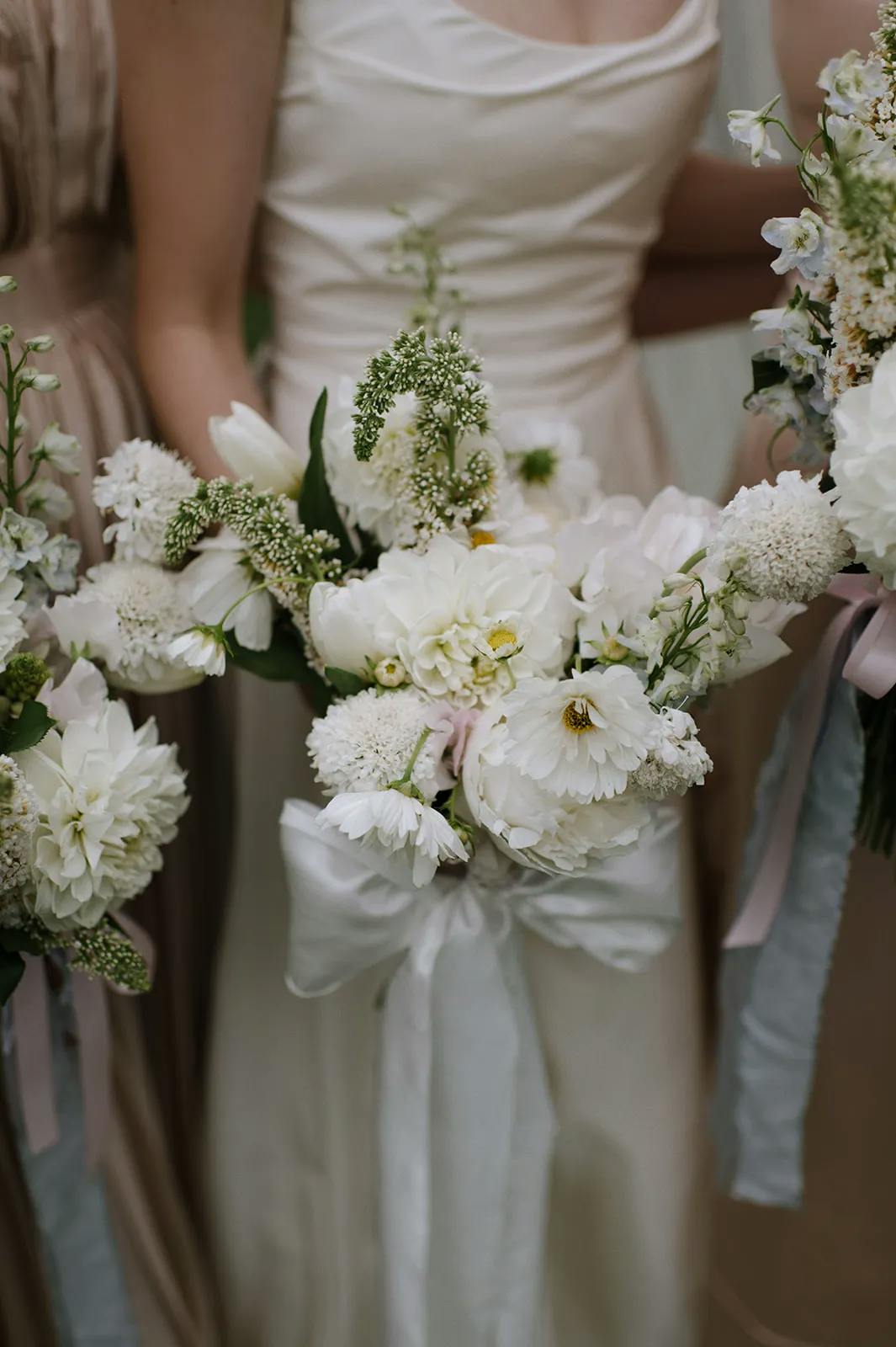 Three people in formal attire are holding floral bouquets. The central bouquet features white flowers with greenery, adorned with a white ribbon. The background is out of focus, highlighting the elegant and delicate arrangement of the flowers.