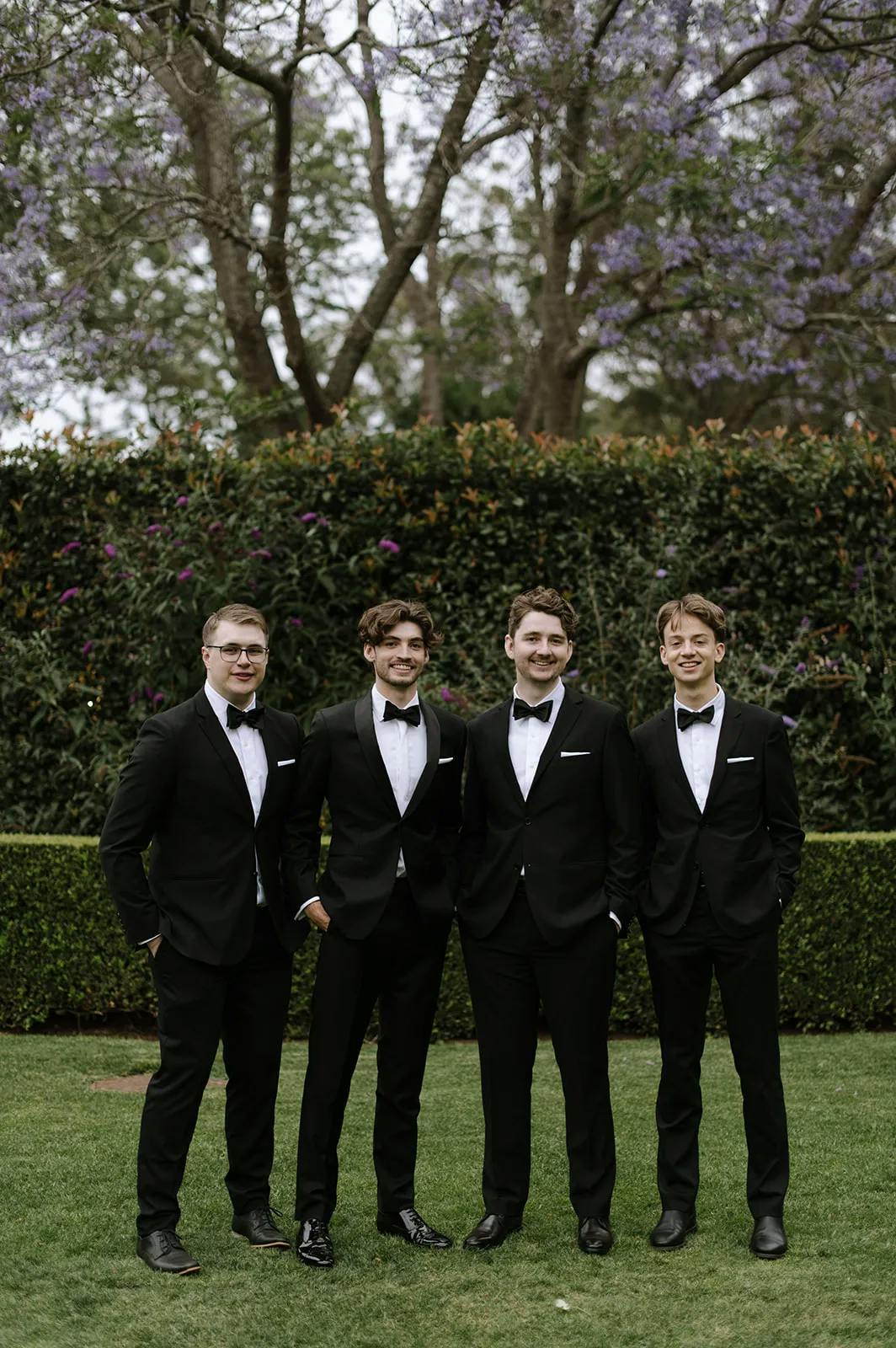 Four men dressed in black tuxedos stand on a lawn with lush greenery and purple flowering trees in the background. They are smiling and standing close to each other, with hands in their pockets or at their sides.