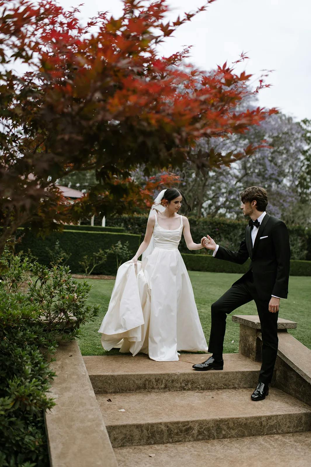 A bride in a white wedding dress holds her skirt as she steps down stone steps, assisted by a groom in a black tuxedo. They are in an outdoor garden with lush greenery and red autumn leaves on a tree in the foreground.