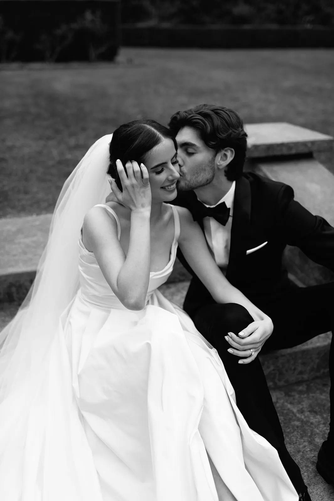 A bride in a white dress and veil sits on steps, smiling, as a groom in a black tuxedo kisses her cheek. Both appear happy and intimate, sharing a tender moment outdoors. The image is in black and white.