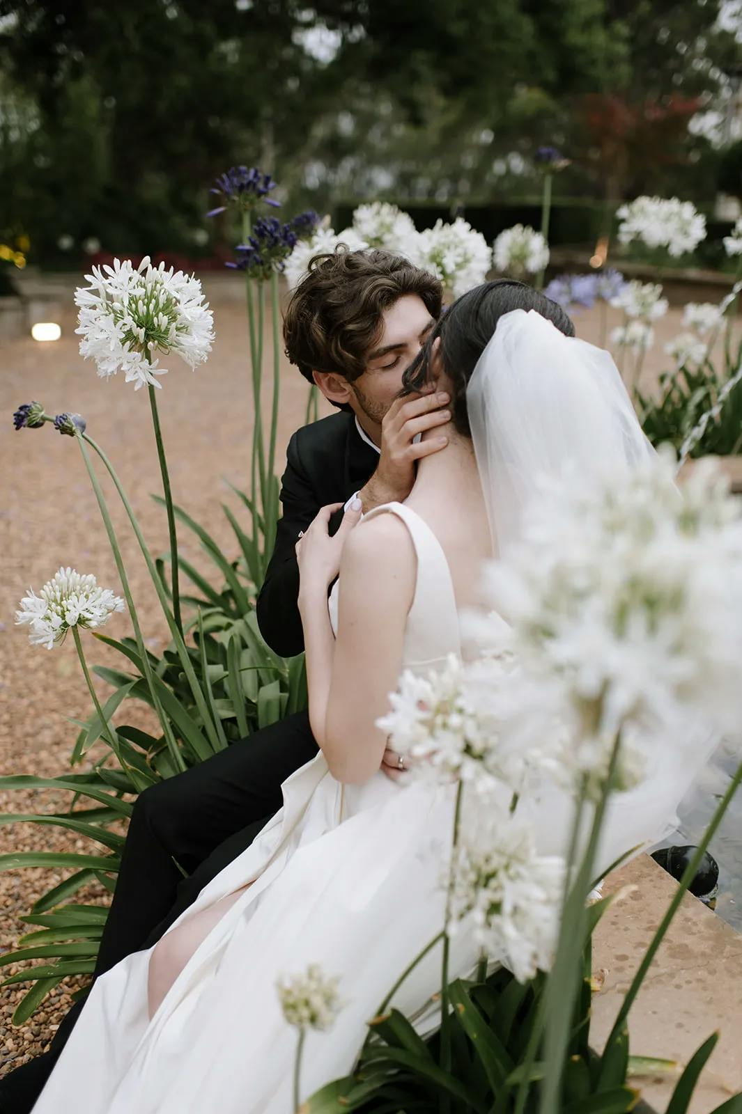 A newly married couple embraces and shares a kiss in a garden, surrounded by white flowers. The groom is dressed in a black suit and the bride is in a white wedding dress with a veil. They are sitting on a stone path, creating a romantic and intimate atmosphere.