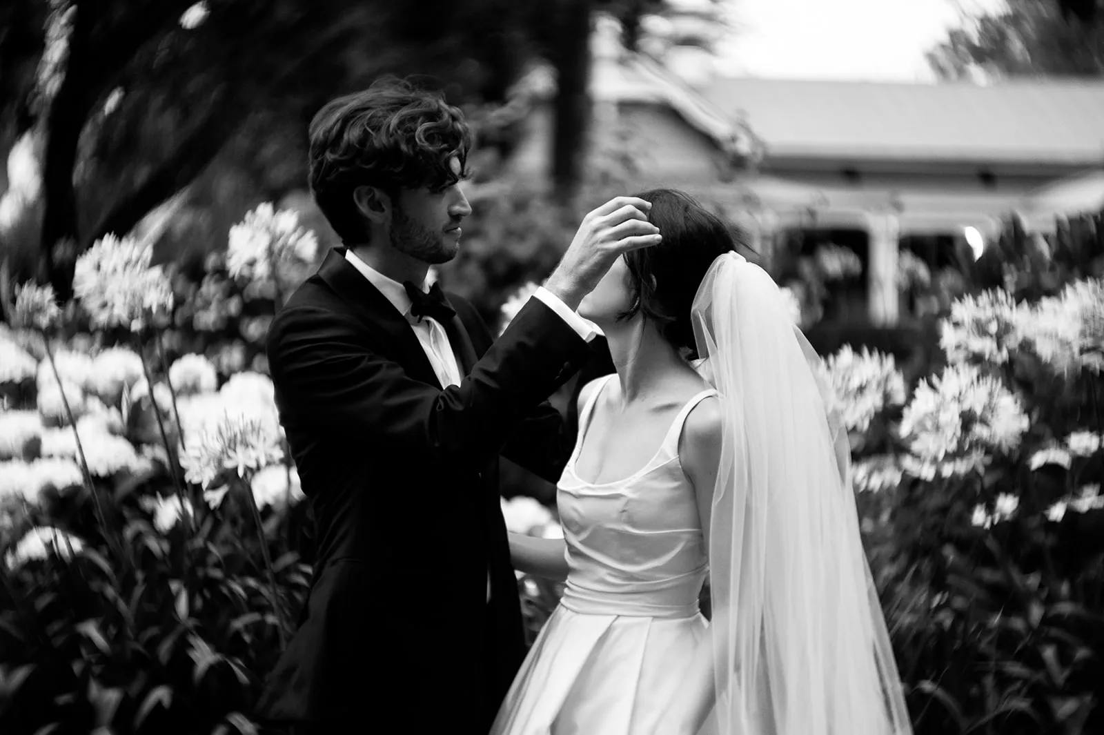 A black-and-white photo of a bride and groom in a garden. The groom, in a dark suit, gently adjusts the bride's hair. The bride, in a white gown with a long veil, looks up at him. Surrounding them are blurred flowers and foliage, with a building faintly visible in the background.