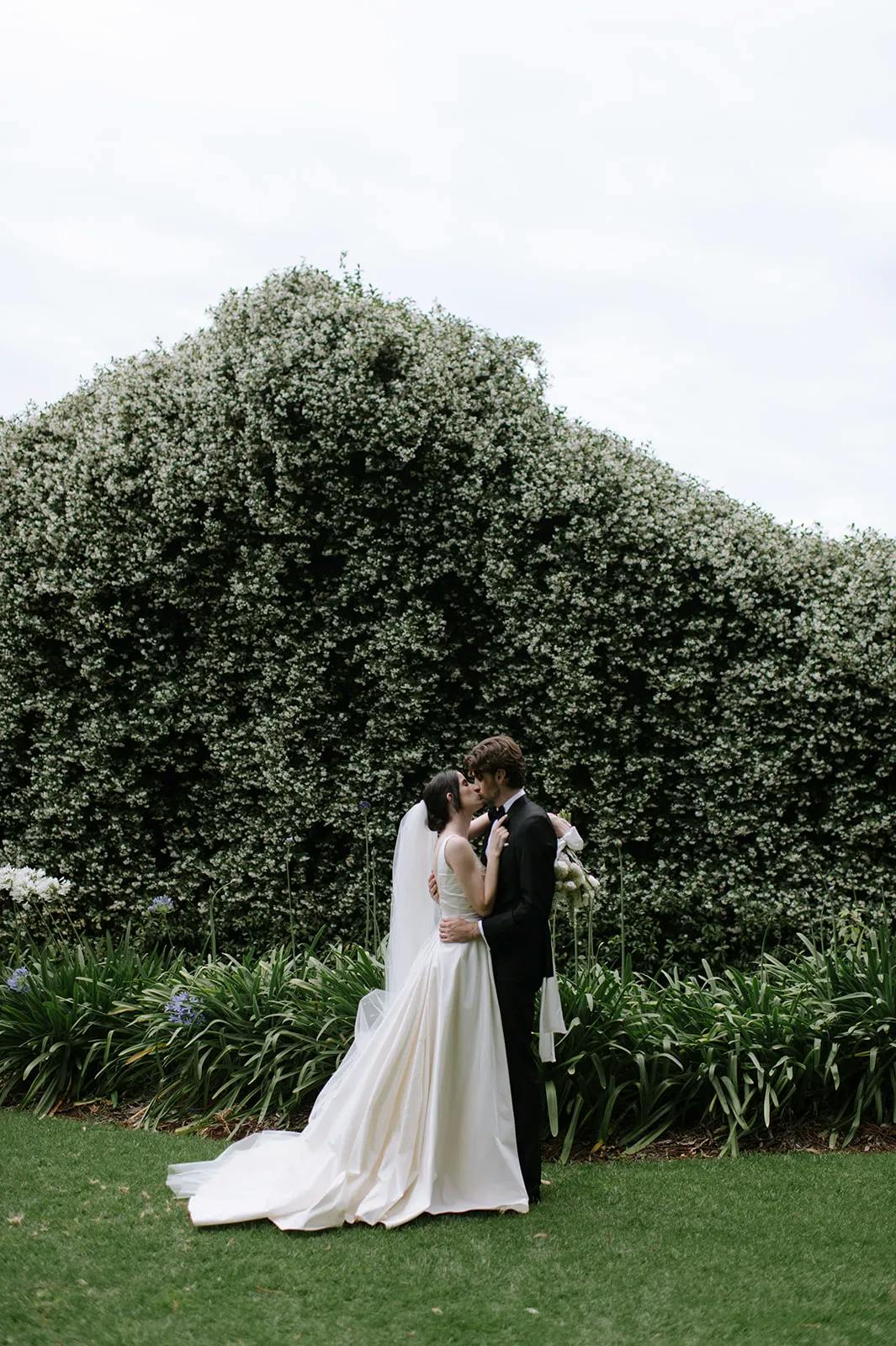 A couple standing closely in wedding attire, sharing a kiss in front of a lush, green hedge covered with white flowers. The bride wears a flowing white gown and veil, while the groom is in a black suit. They are surrounded by greenery and flowers.