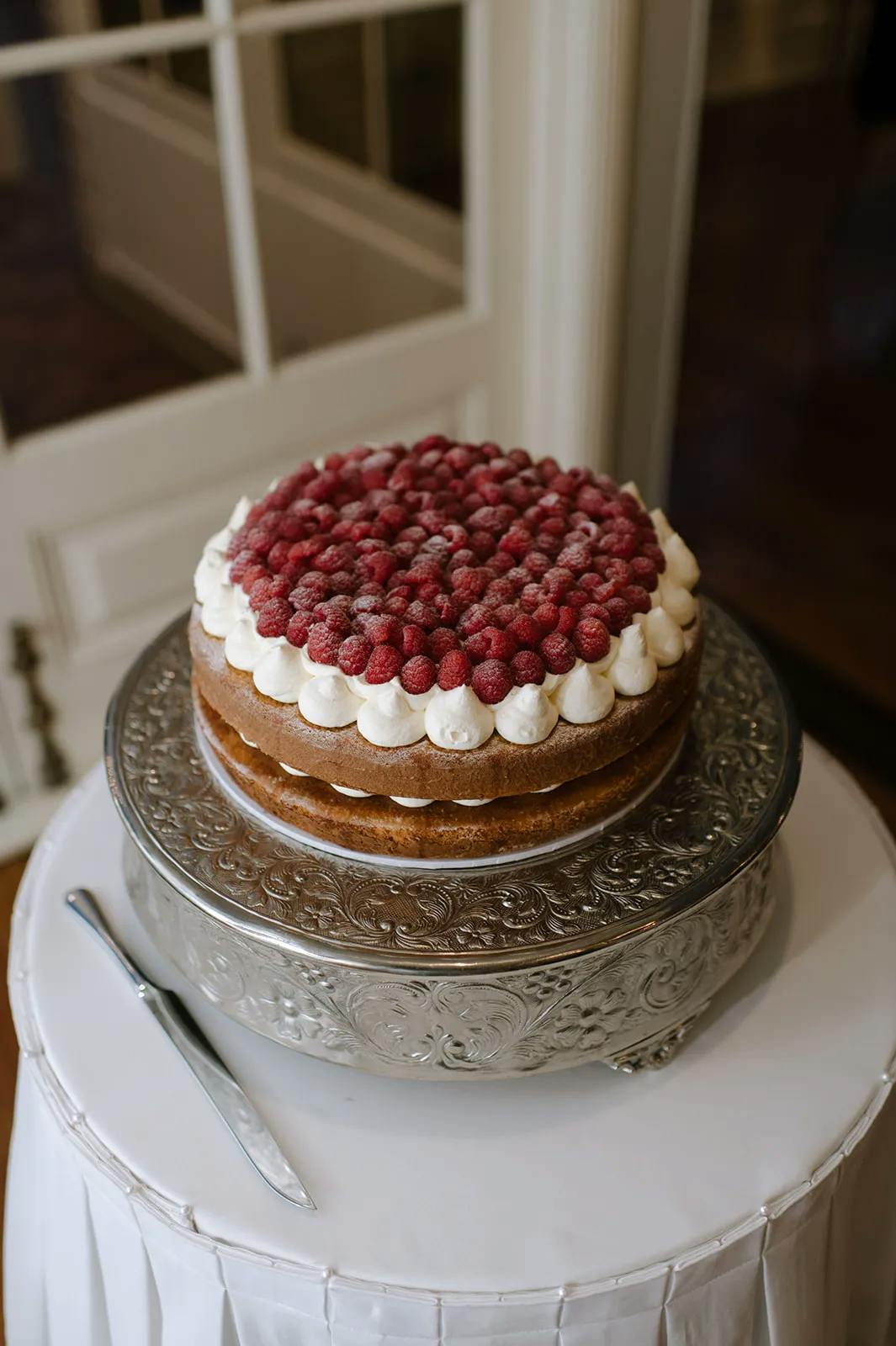 A round layer cake decorated with raspberries and cream sits on an ornate silver cake stand. There's a knife placed on a white tablecloth beside the cake. The background shows a wooden floor and a white door with glass panes.