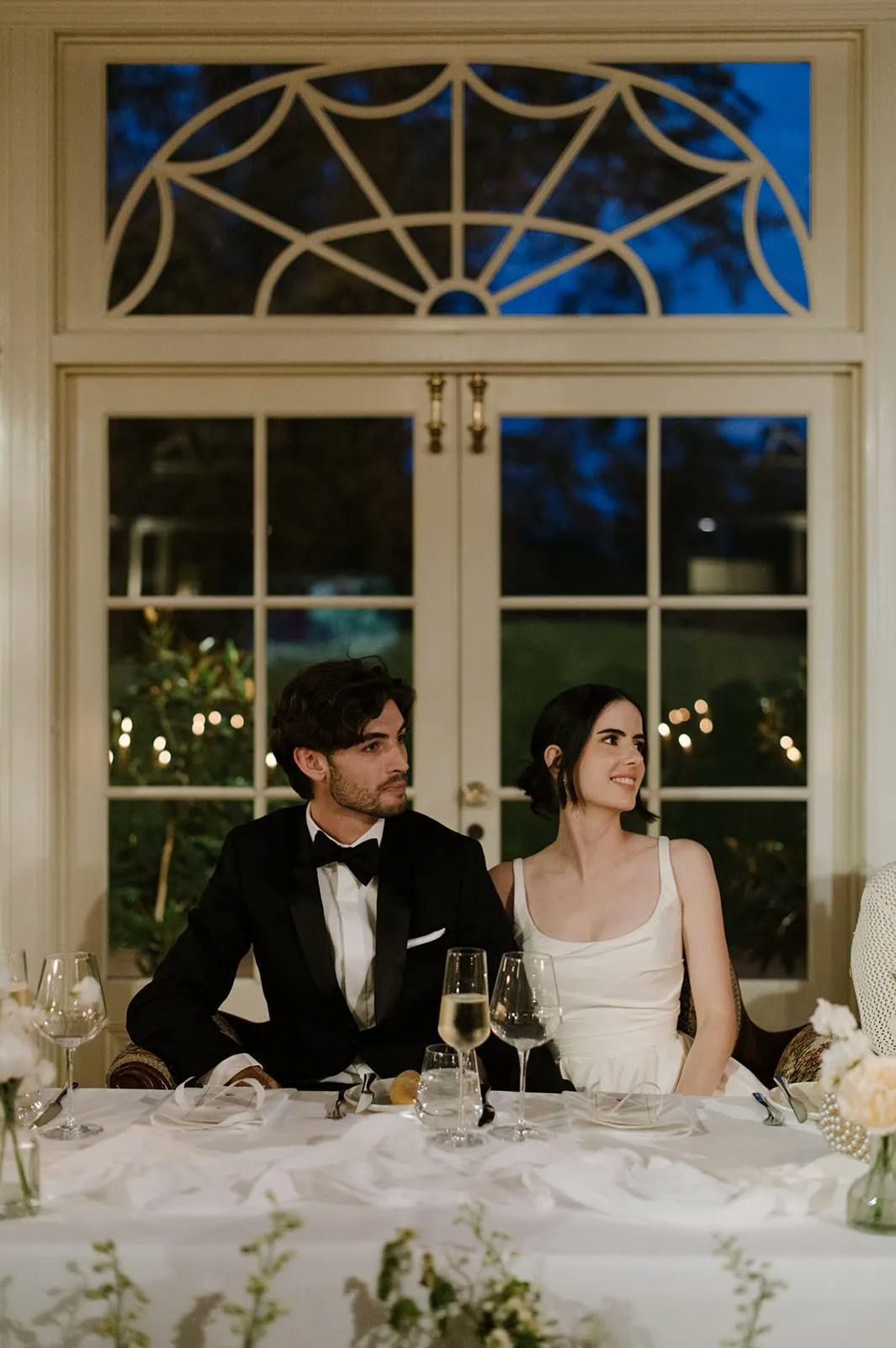 A man in a black tuxedo and a woman in a white dress sit at a table, smiling, during an evening event. They are inside a room with a large window and decorative architecture behind them. The table is set with glassware and floral arrangements.
