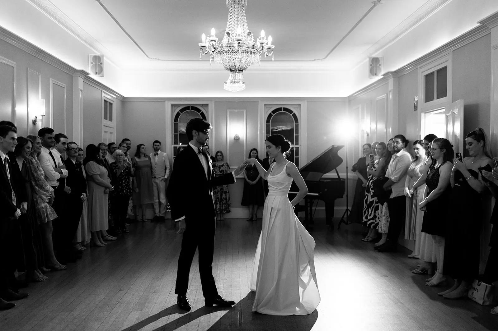A black-and-white photo capturing a couple's first dance at their wedding. They are dressed in formal attire, with the bride in a strapless gown and the groom in a suit. They are surrounded by guests watching and standing in a spacious room with a chandelier and a grand piano.