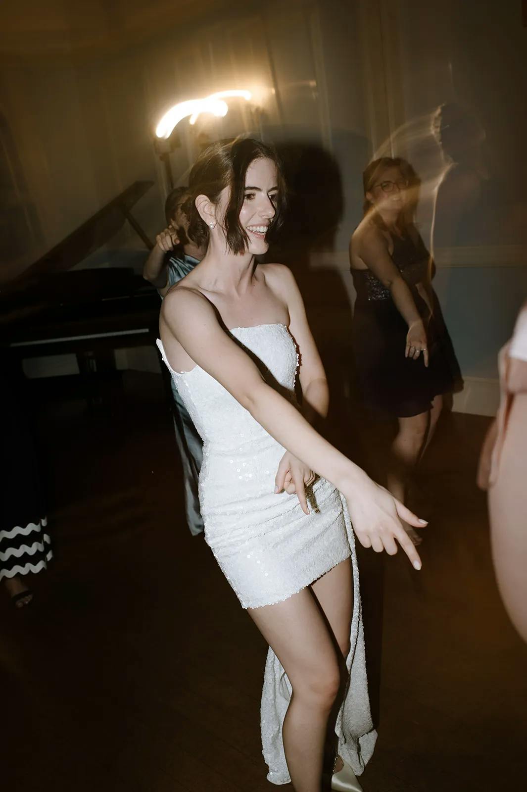 A woman in a white, sequined strapless dress dances joyfully at an indoor event. She is smiling widely and seemingly in motion with one arm extended. Some blurred people are in the background, suggesting movement and lively activity.