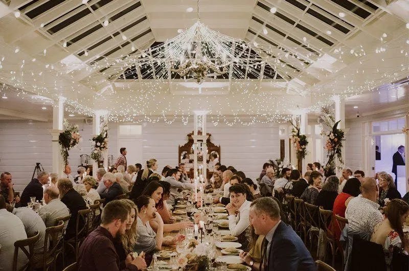A large, elegantly decorated hall with string lights hanging from the ceiling, hosting a dinner event. Numerous people are seated at long, banquet-style tables covered with plates, glasses, and floral centerpieces. The atmosphere is festive and lively.