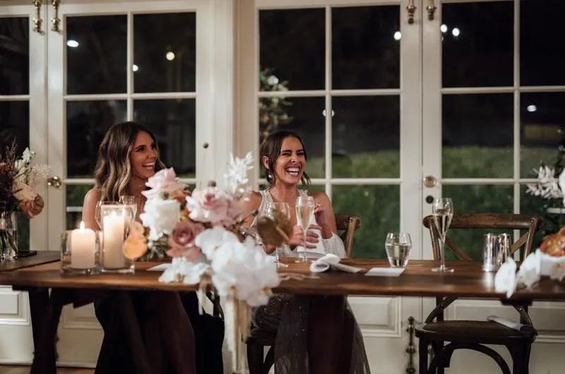 Two women dressed in formal attire sit at a beautifully decorated table, laughing and holding glasses. The table is adorned with elegant flower arrangements and lit candles. Behind them are large windows showing a dark outdoor scene. The atmosphere is warm and festive.