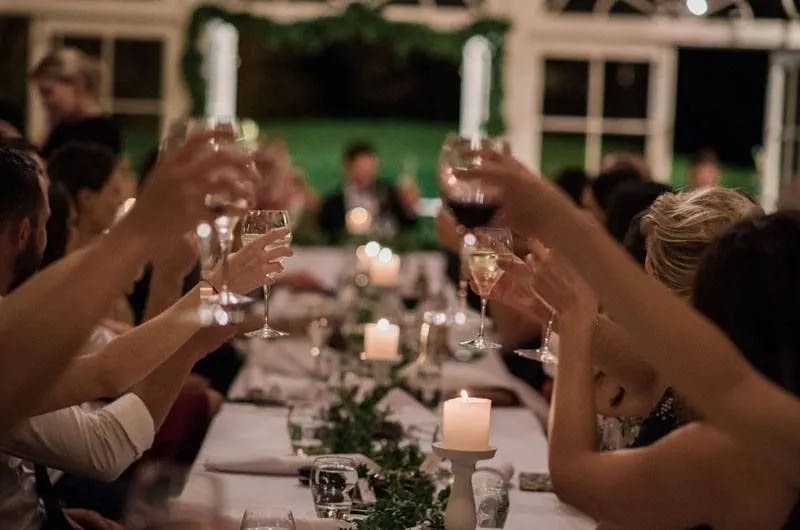 A group of people seated at a decorated, candlelit table raise their glasses for a toast during a formal gathering or celebration. Green foliage and white candles line the center of the table, and the background shows an open room with large windows.