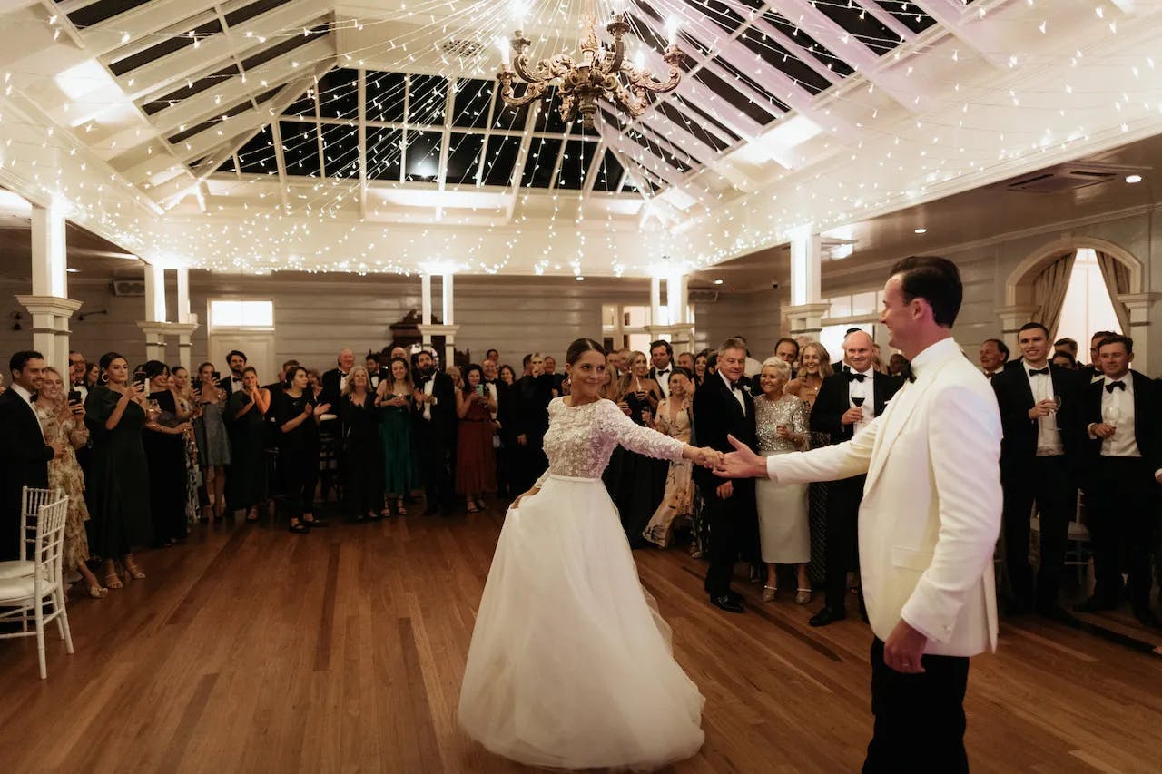 A bride and groom share their first dance under a ceiling adorned with fairy lights and a chandelier. The bride wears a white gown, while the groom dons a white tuxedo jacket. Guests in formal attire surround them, smiling and capturing the moment with their phones.