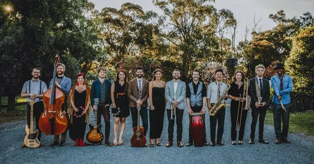 A diverse group of 13 musicians stands outdoors in a line, each holding their instruments, including guitars, a double bass, keyboard, saxophone, trumpet, trombone, violin, and conga drums. They are dressed in formal and semi-formal attire, with trees in the background.