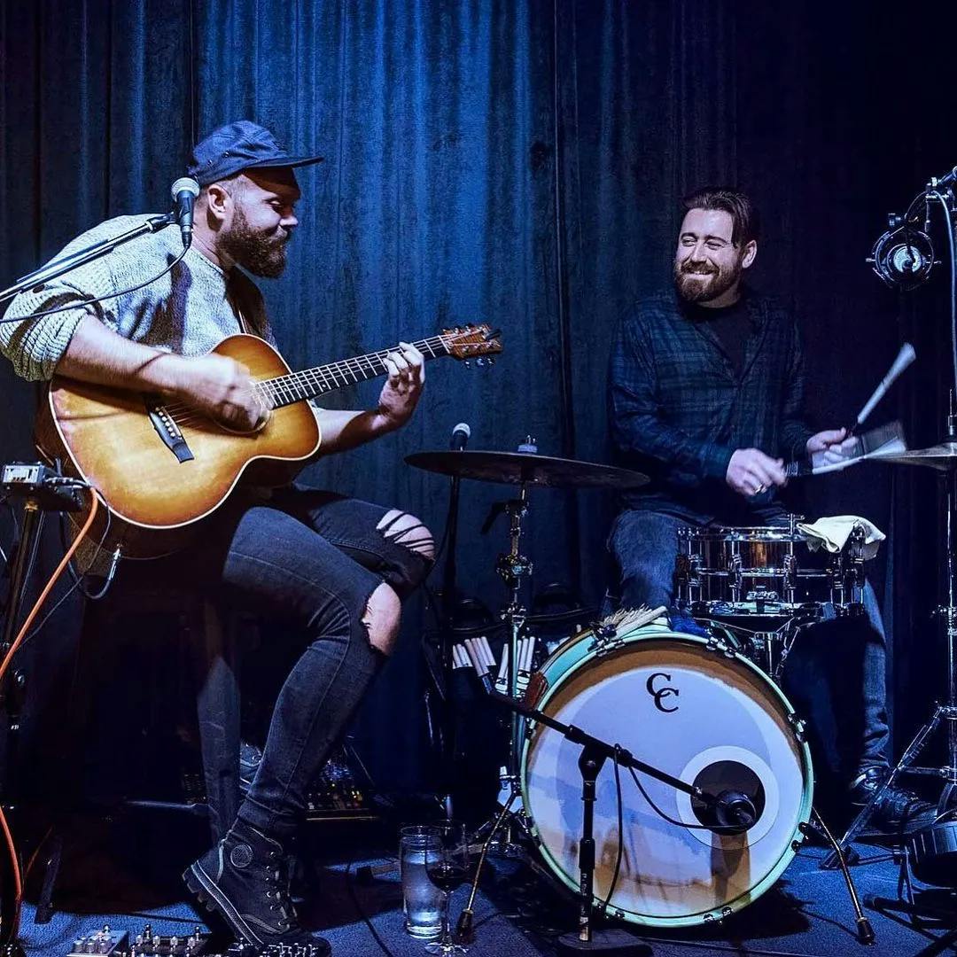 Two musicians performing on a dimly lit stage. The one on the left is playing an acoustic guitar and singing, while the bearded musician on the right is playing the drums and smiling. Both appear to be enjoying the music. A blue curtain serves as the backdrop.