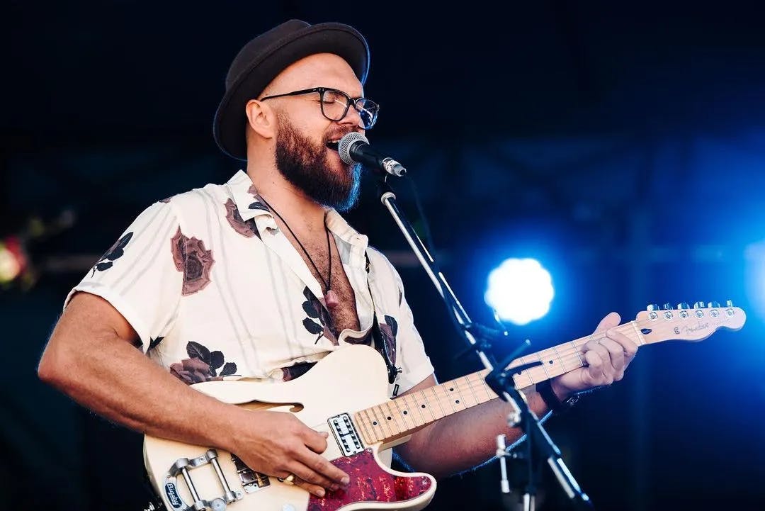 A bearded man wearing a hat and glasses plays an electric guitar and sings into a microphone on stage. He is dressed in a light-colored shirt with a floral pattern. The background is dark with bright stage lights shining.