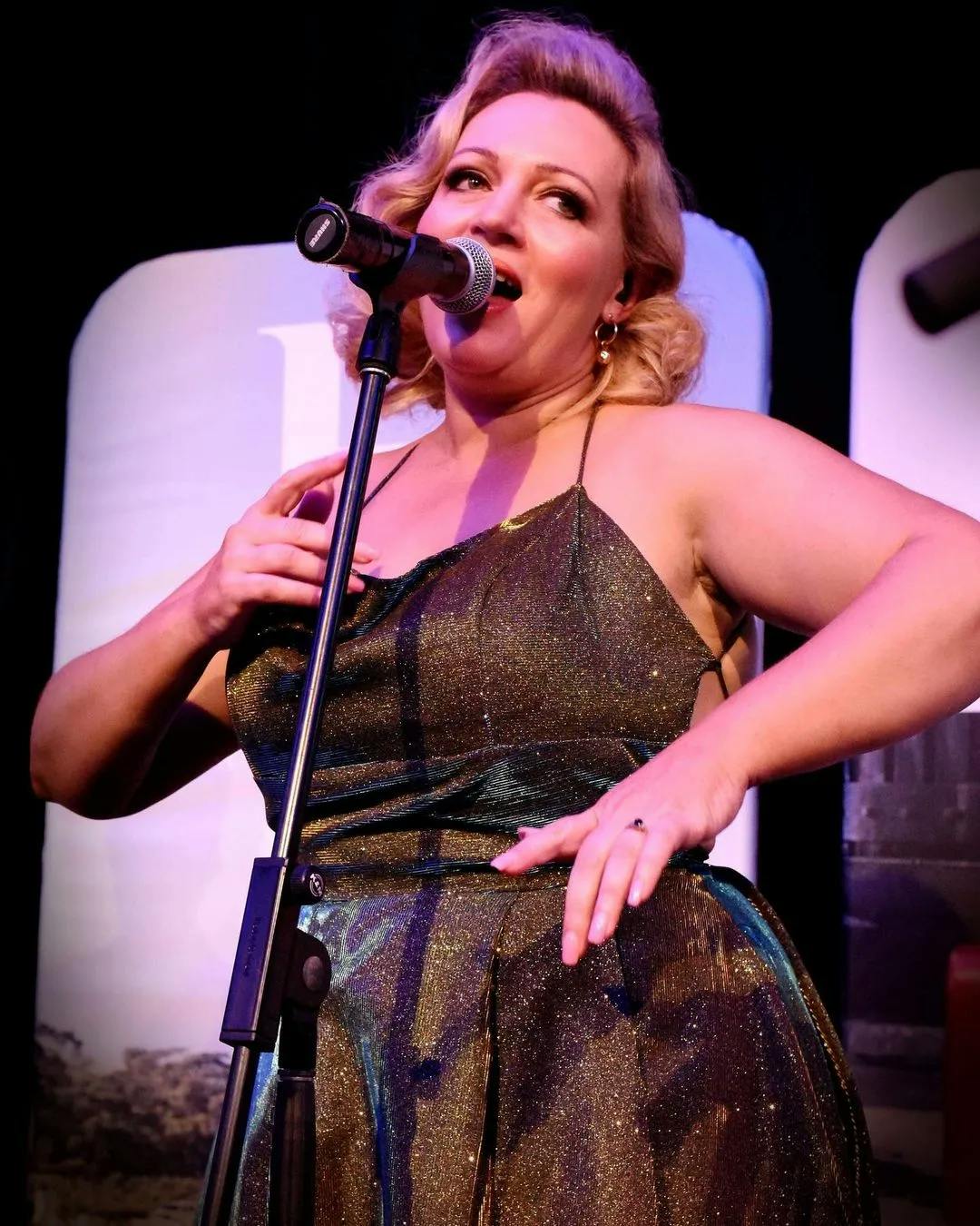 A woman with blonde hair performs on stage, singing into a microphone. She is wearing a shimmery, metallic dress and appears to be in the middle of a dynamic performance, her hand elegantly resting on her chest. The background is dark with some illuminated signage.