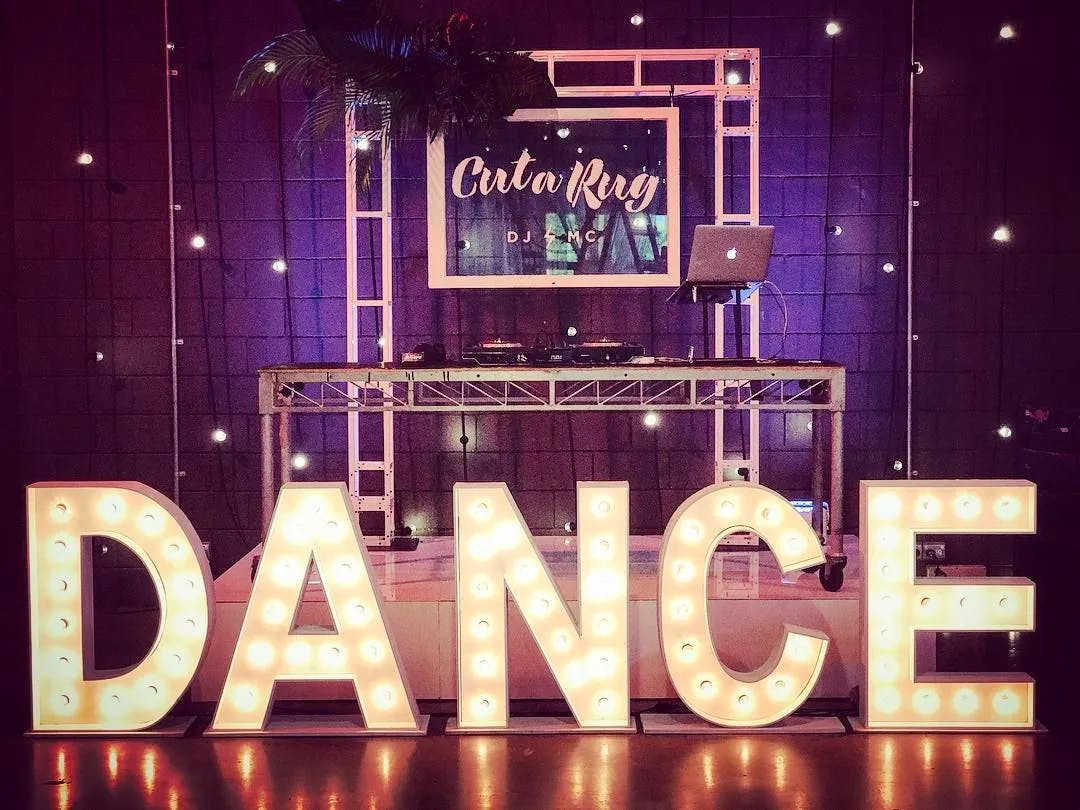 A brightly lit dance floor setup with a large, illuminated sign spelling "DANCE" in front of a DJ booth. The backdrop features a framed sign with "Cut a Rug DJ - MC" and string lights creating a festive atmosphere. A laptop and DJ equipment are on the booth.