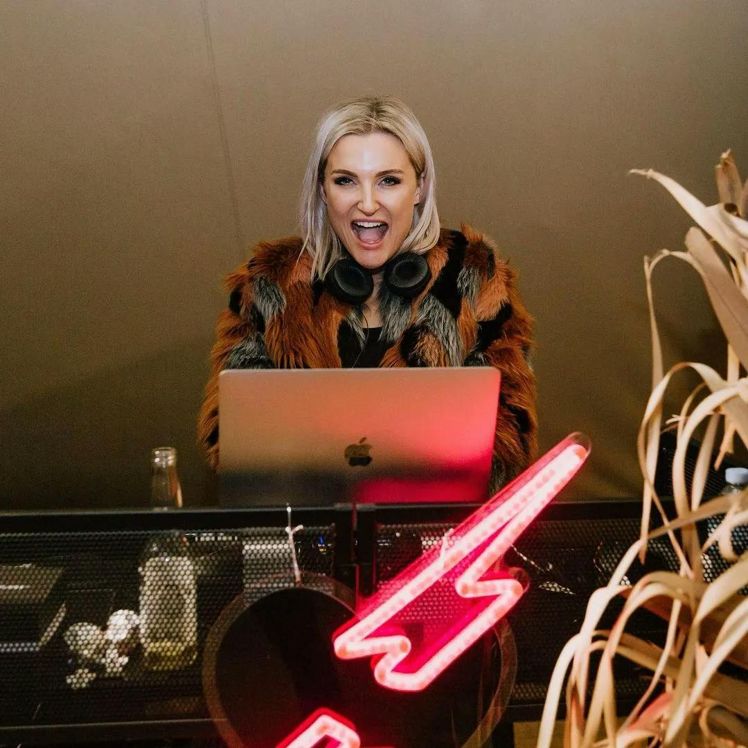 A person with blonde hair, wearing a furry jacket with black and orange patterns, stands behind a laptop with headphones around their neck. The setup is on a surface with a neon lightning bolt sign and some decorative plants in the foreground.