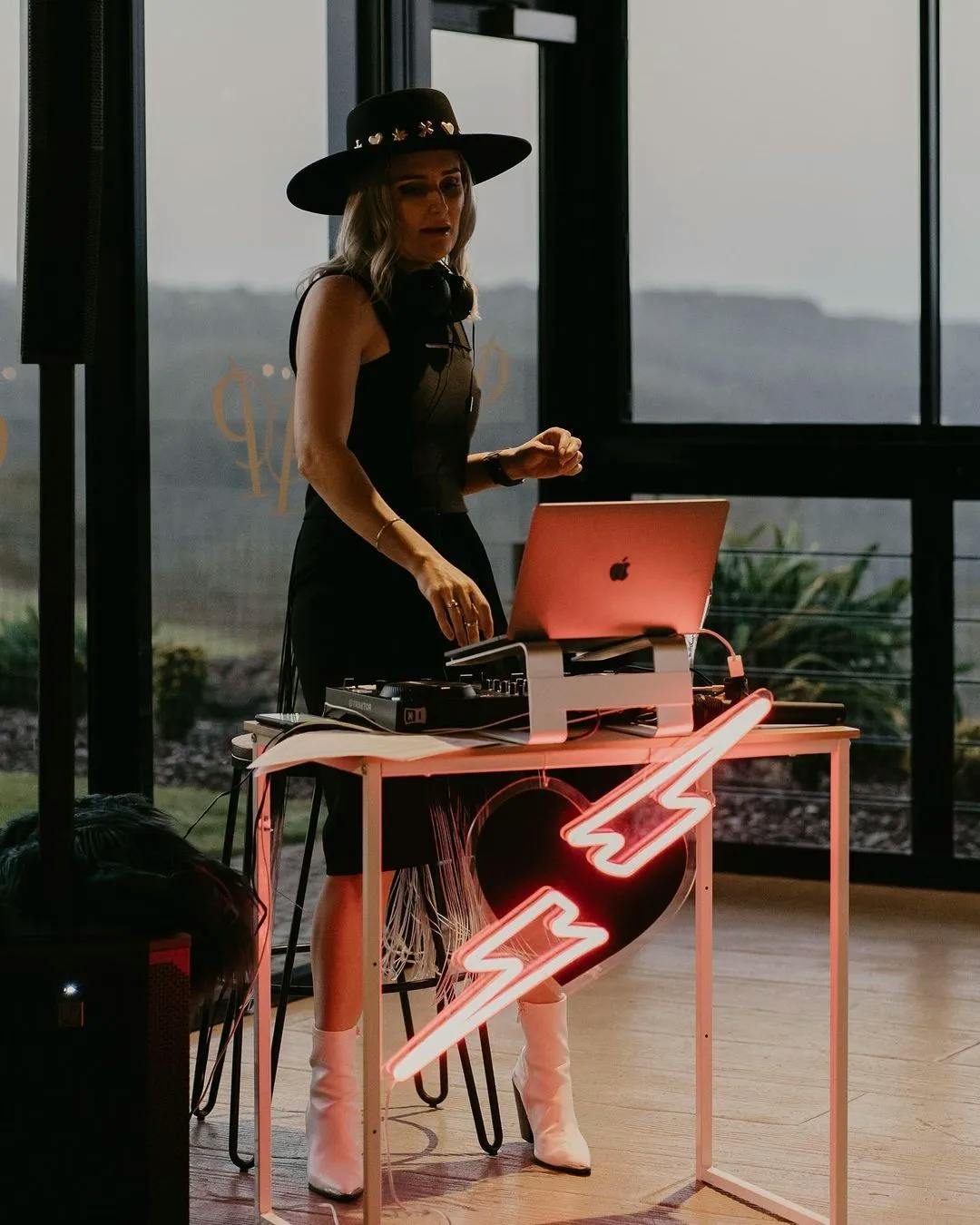 A DJ wearing a black dress, wide-brimmed hat, and white boots stands behind a table with a laptop and DJ equipment. The table is decorated with a neon light shaped like lightning bolts. The background shows large windows overlooking a landscape.