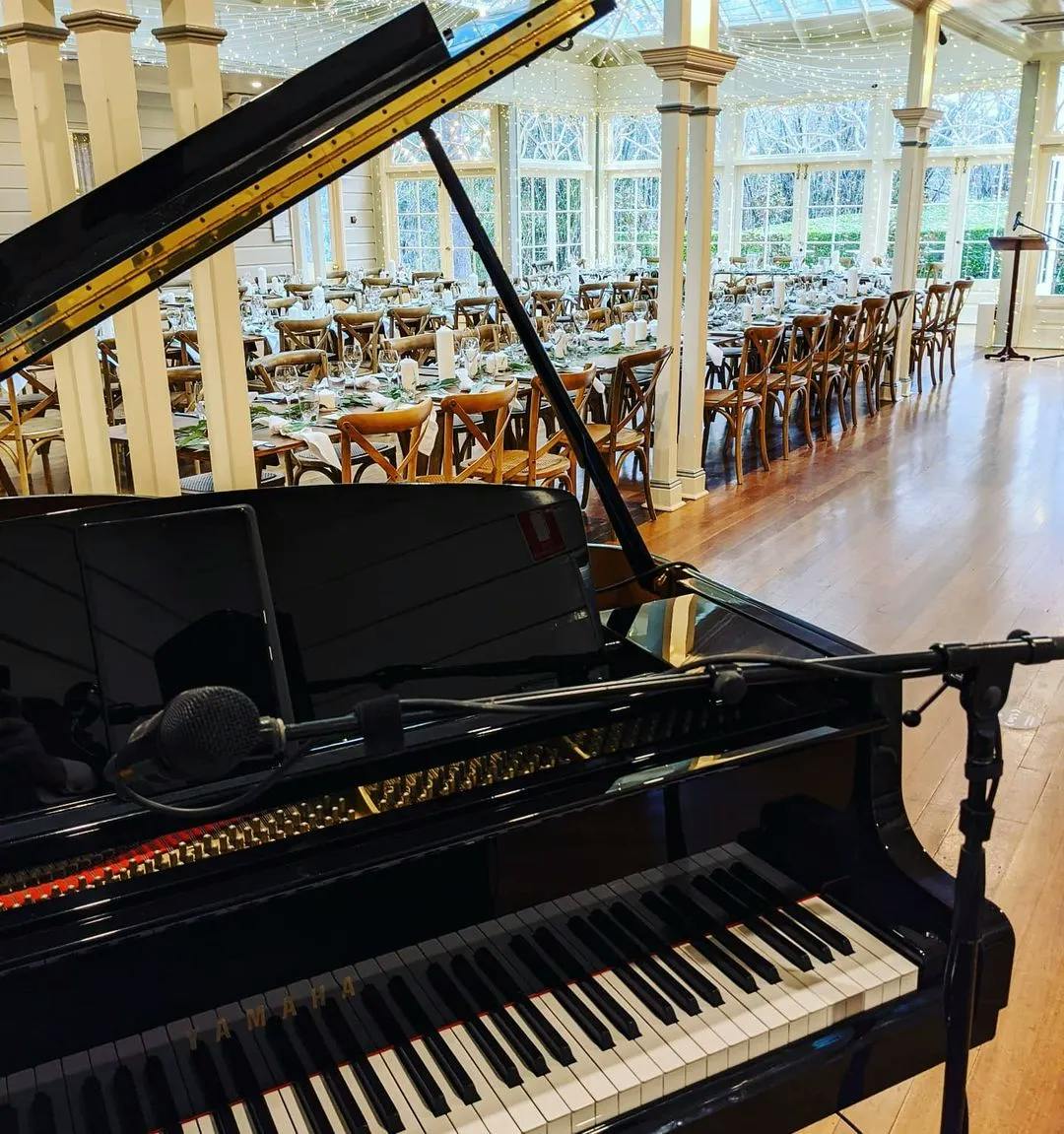 A grand piano with a microphone setup is placed in the foreground of a spacious hall with wooden floors. The hall is decorated with long banquet tables and wooden chairs, set for an event, and large windows letting in natural light.