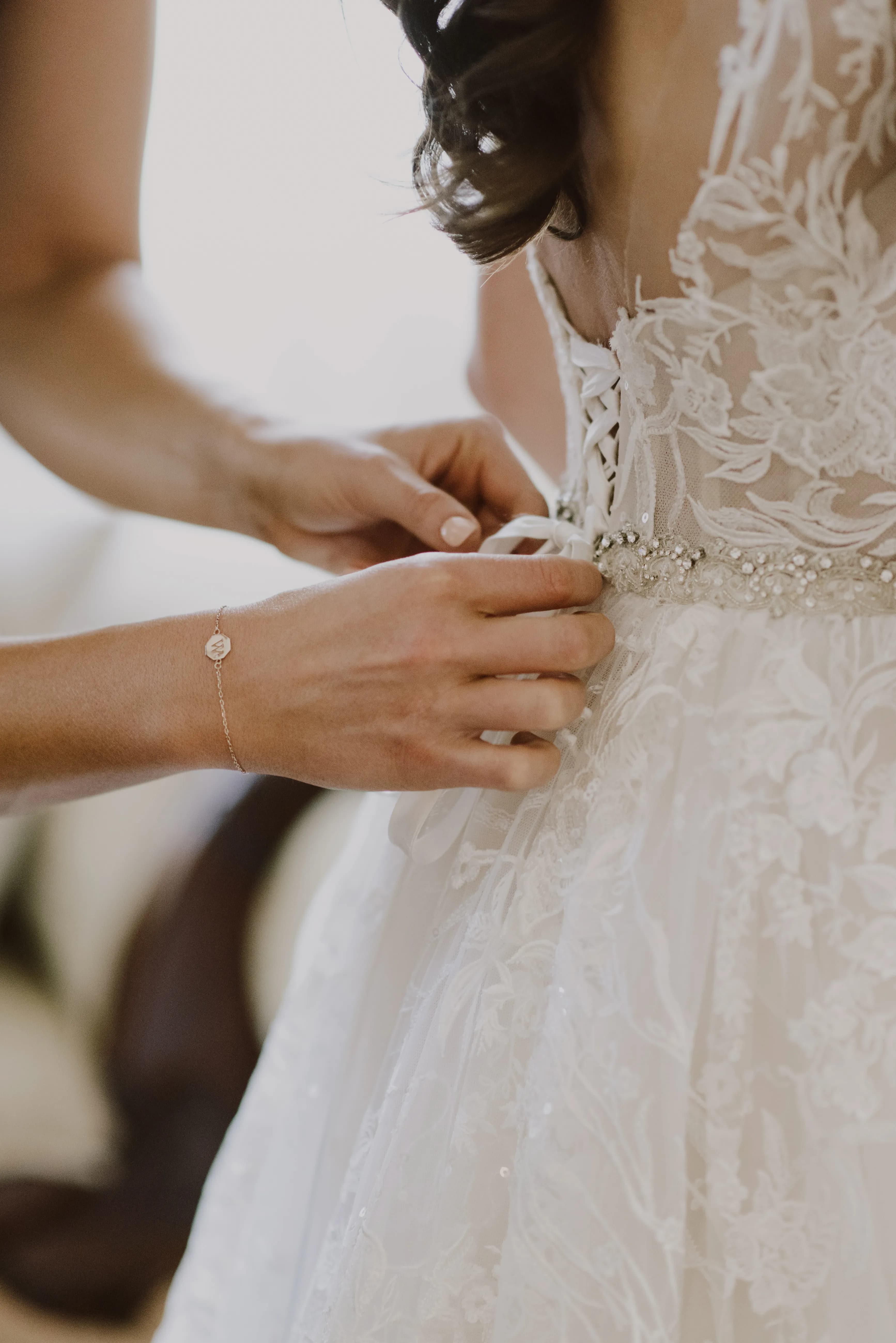 A close-up of a bride in an intricate lace wedding dress being fastened at the back by someone. The bride's dress features detailed floral embroidery and a bead-embellished belt. The person fastening the dress wears a delicate bracelet with a small charm.