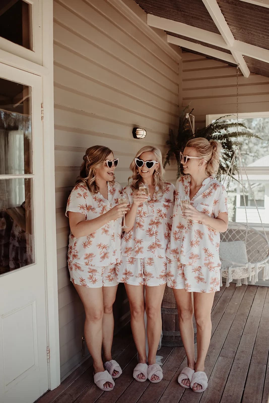 Three women stand outside on a wooden porch, wearing matching pajamas with a white and pink floral pattern. They are wearing sunglasses and holding glasses of drinks, smiling and talking to each other. There is a hanging chair and potted plant in the background.
