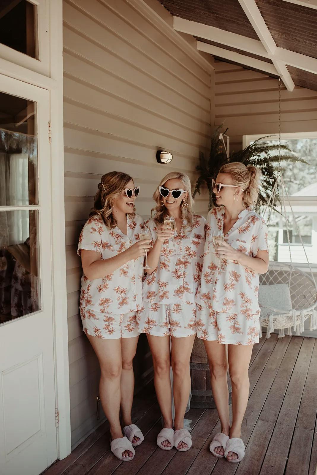 Three women stand outside on a wooden porch, wearing matching pajamas with a white and pink floral pattern. They are wearing sunglasses and holding glasses of drinks, smiling and talking to each other. There is a hanging chair and potted plant in the background.