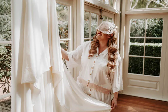 A woman wearing a white sleep mask and light-colored pajamas smiles as she touches the fabric of a wedding dress hanging by a window. The room is bright, with large windows revealing a lush green garden outside.