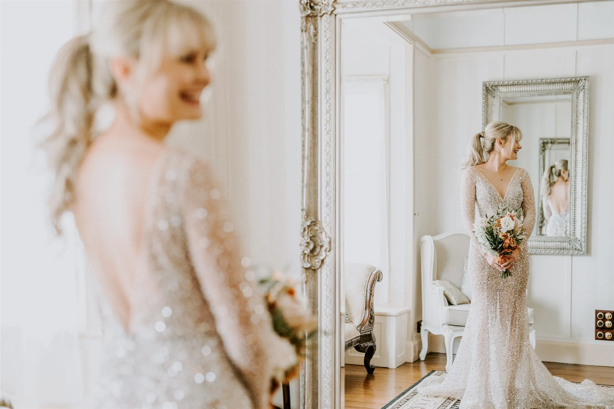 A bride with blonde hair in a ponytail is wearing a sparkling, long-sleeved wedding gown with a deep V-neck. She stands in front of a large, ornate mirror holding a bouquet of flowers, smiling at her reflection in a bright and elegant room with white decor.