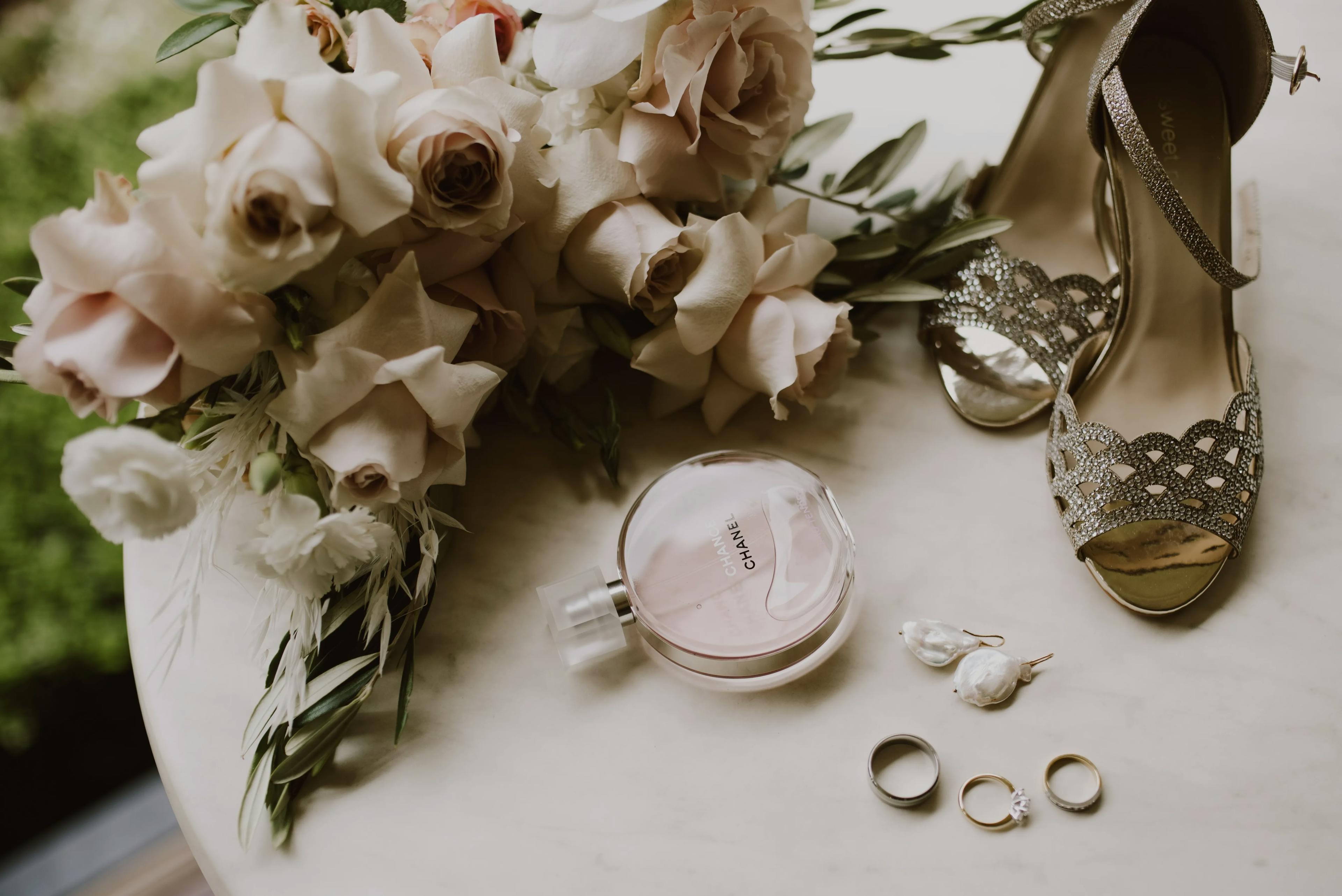 A bouquet of white and blush roses lies on a table next to a bottle of Chanel perfume, a pair of elegant silver high heels, two rings, a set of drop earrings, and some greenery. The items are displayed on a light-colored surface.