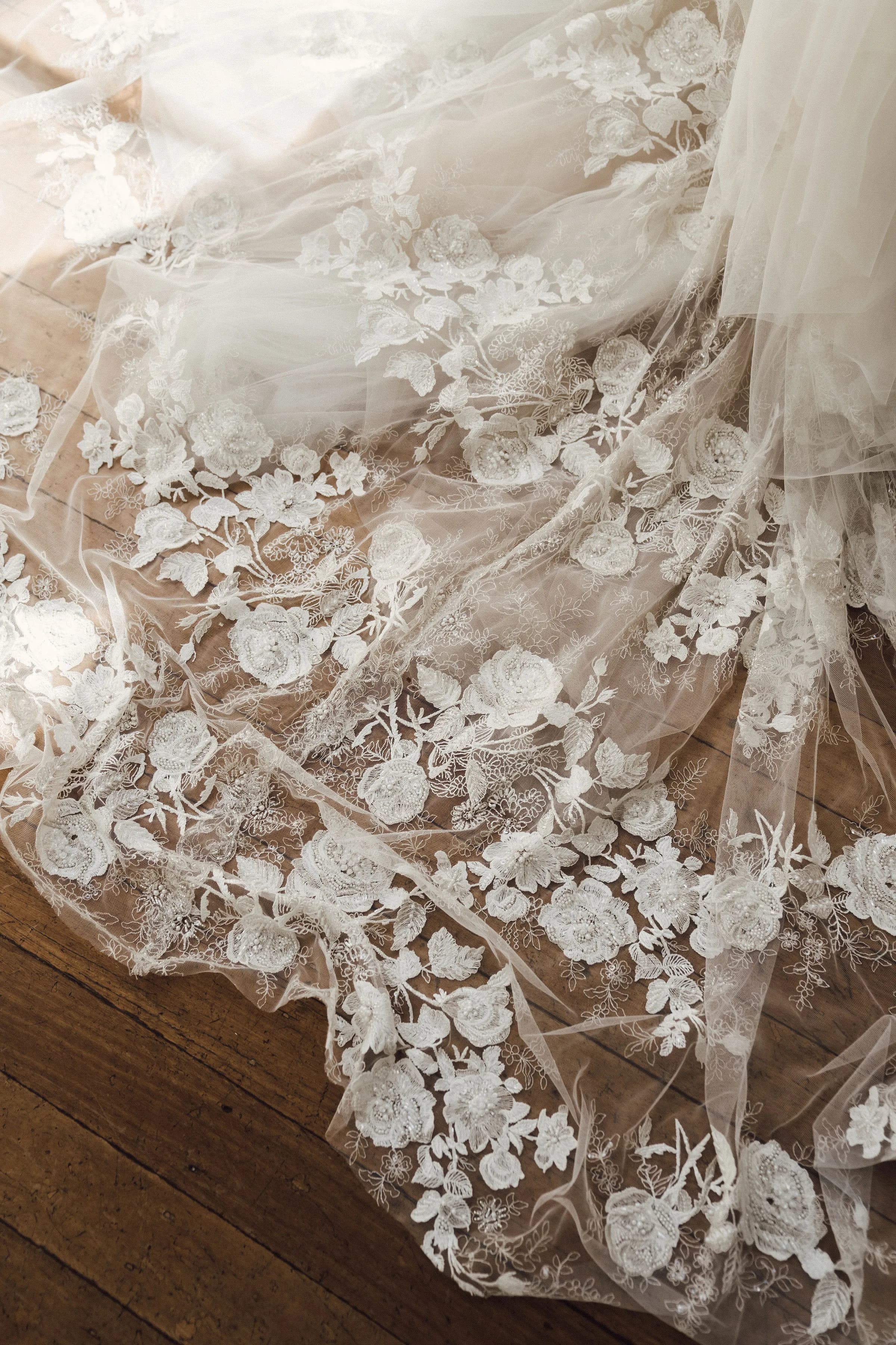 A close-up of a wedding dress train spread out over a wooden floor. The delicate fabric is adorned with intricate white lace floral patterns, featuring roses and other flowers, creating an elegant and romantic appearance.