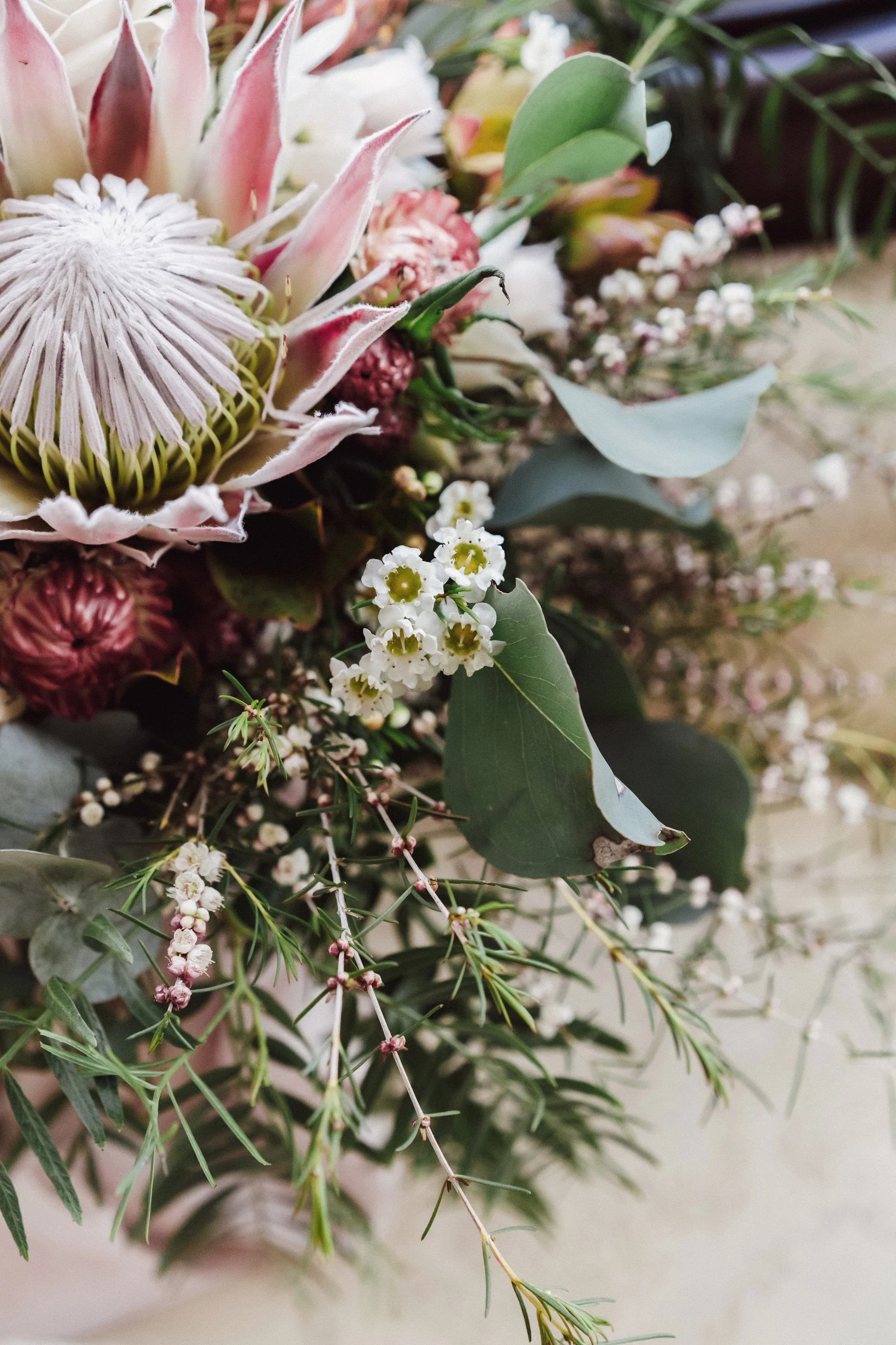 A detailed close-up of a lush floral arrangement featuring a prominent pink protea flower, surrounded by small white blossoms, green eucalyptus leaves, and other assorted foliage. The combination of textures creates a natural and vibrant appearance.