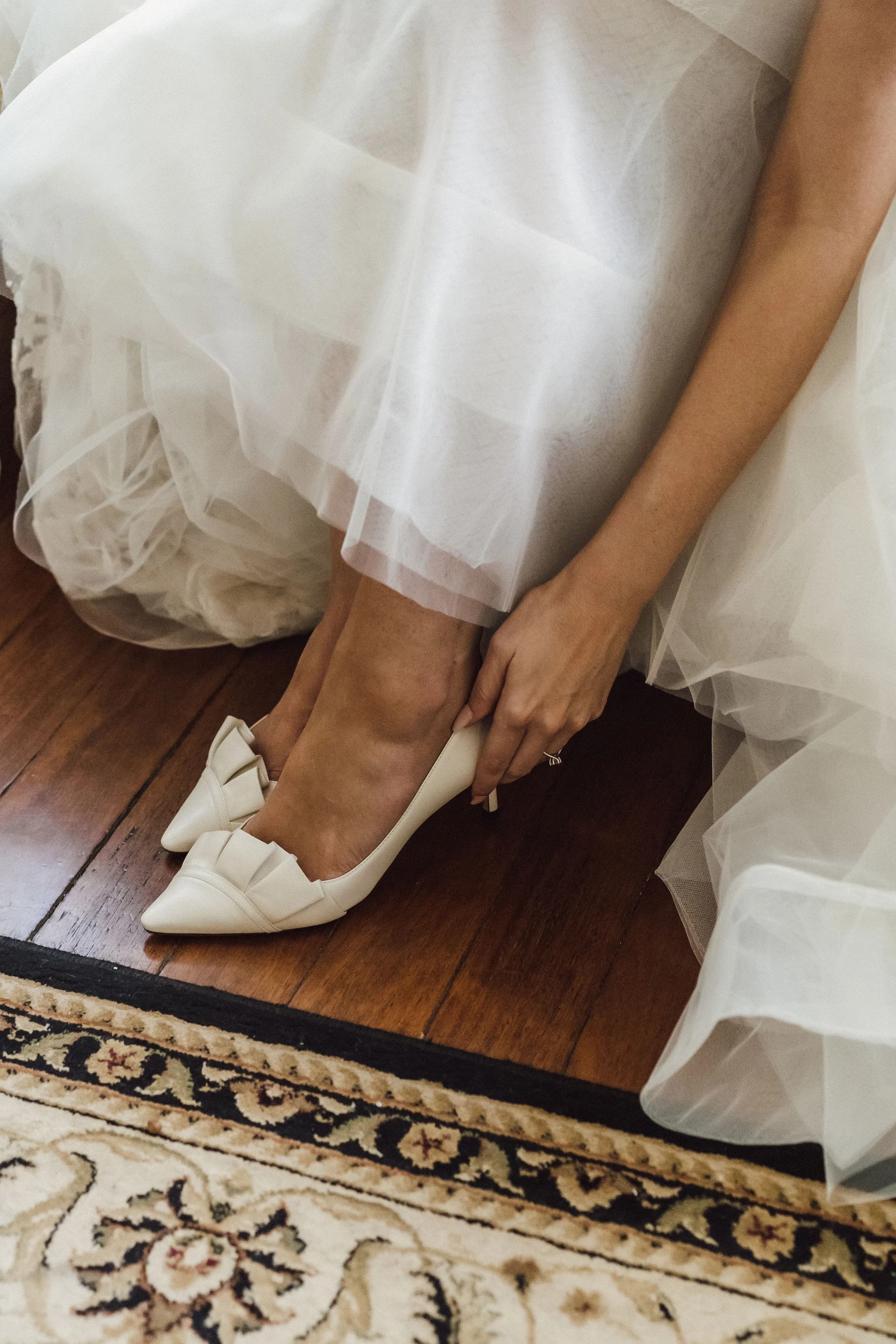 A person dressed in a white bridal gown is adjusting their white high-heeled shoes adorned with bows, seated on a wooden floor next to a patterned rug. Only the lower half of their body is visible in the image.