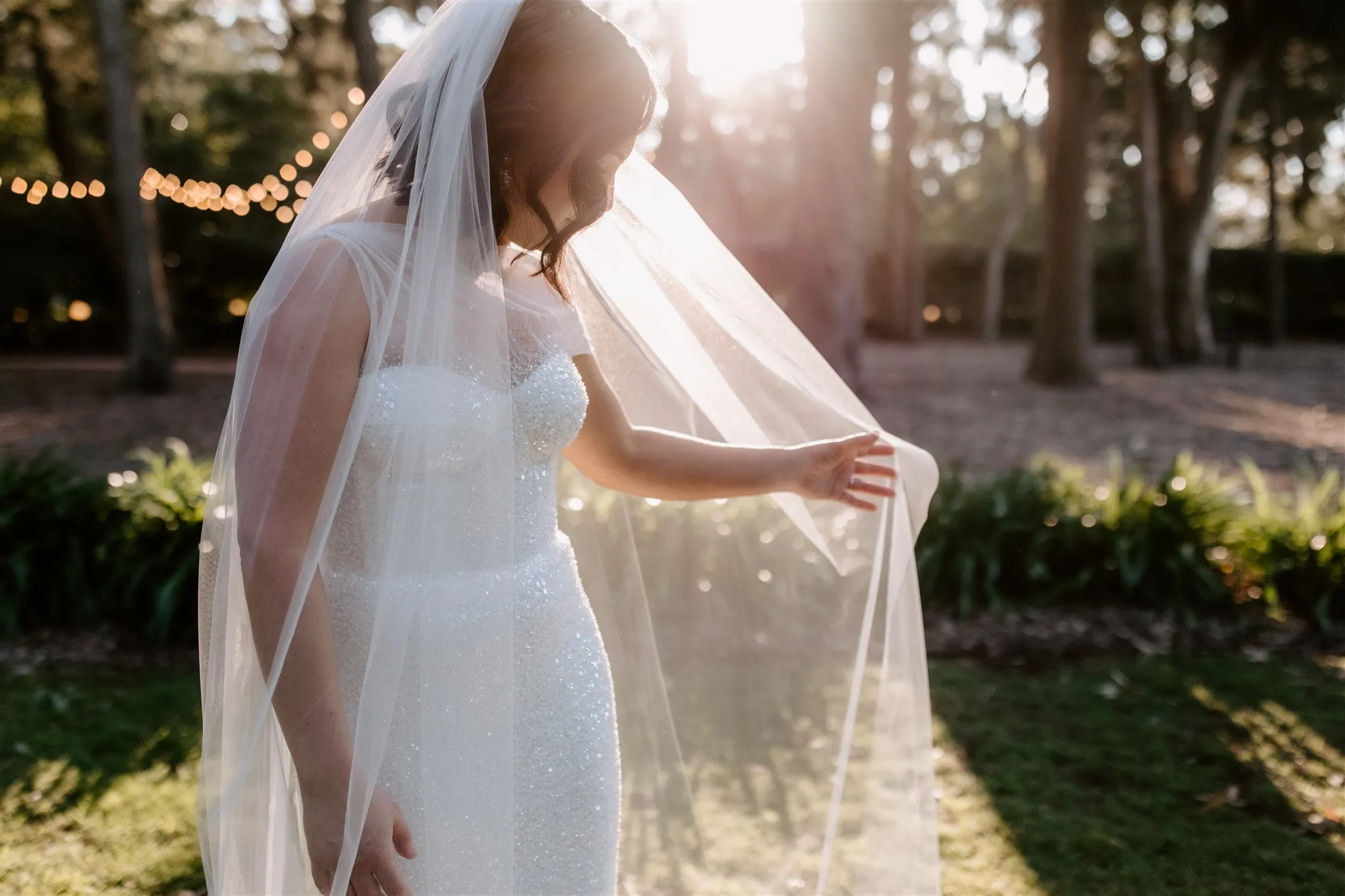 A bride in a sparkly white wedding dress stands in an outdoor setting with sunlight streaming through the trees. She is holding her sheer veil with one hand, and the background features string lights, greenery, and soft natural light.
