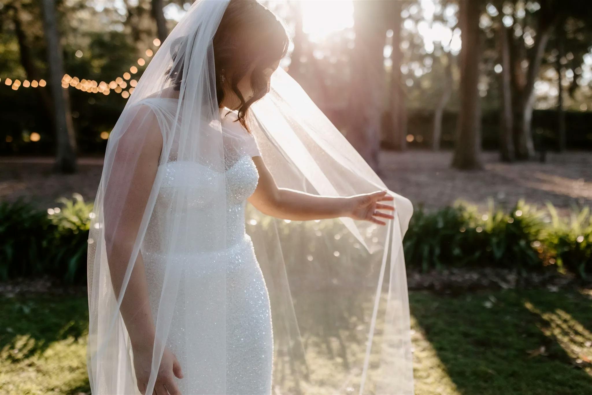 A bride in a sparkly white wedding dress stands in an outdoor setting with sunlight streaming through the trees. She is holding her sheer veil with one hand, and the background features string lights, greenery, and soft natural light.