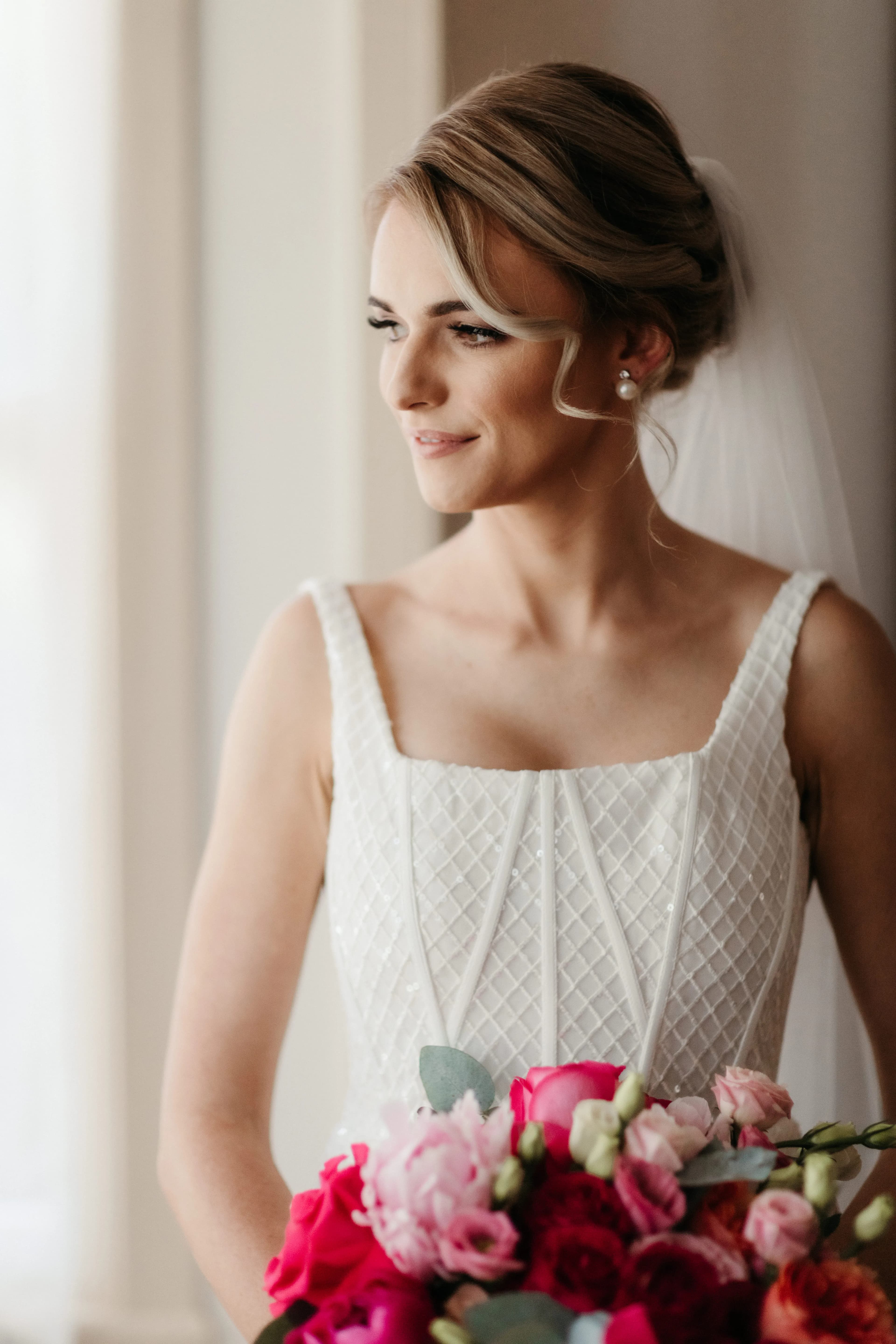 A bride stands near a window, gazing outside with a gentle smile. She wears a sleeveless, white, textured wedding gown and holds a vibrant bouquet of pink, red, and white flowers. Her hair is styled elegantly and is adorned with a veil.