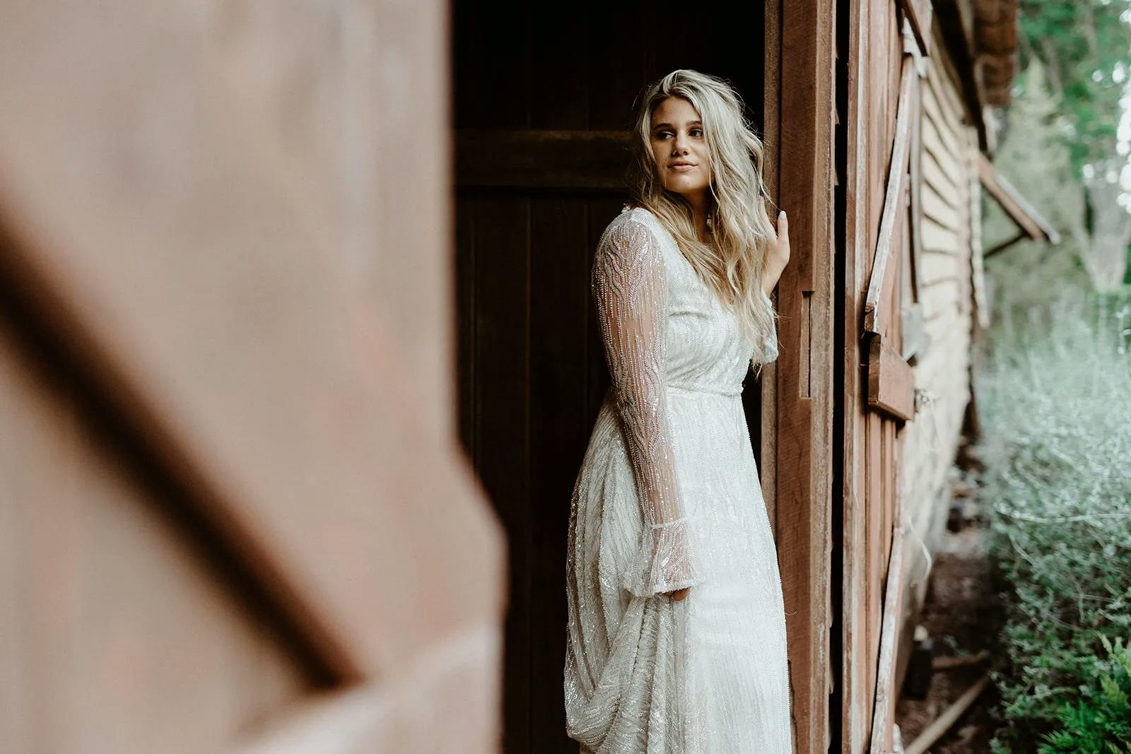 A woman in a white, long-sleeve dress with textured fabric stands in a partially open doorway of a rustic wooden building, looking off into the distance with a relaxed, thoughtful expression. She has long, wavy blonde hair and there is greenery in the background.