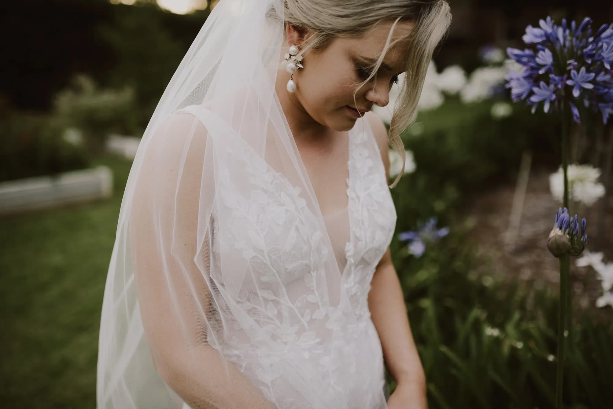 A bride in a white lace wedding dress stands outdoors in front of purple flowers. She is looking down and wearing a sheer veil and pearl earrings. The background is green and lush, suggesting a garden setting.