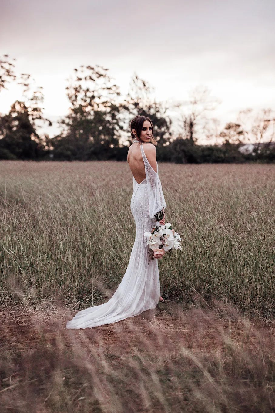 A bride in a backless white wedding dress stands in a field of tall grass, looking over her shoulder. She holds a bouquet of white and pink flowers. Trees are visible in the background under a softly lit, cloudy sky at dusk.