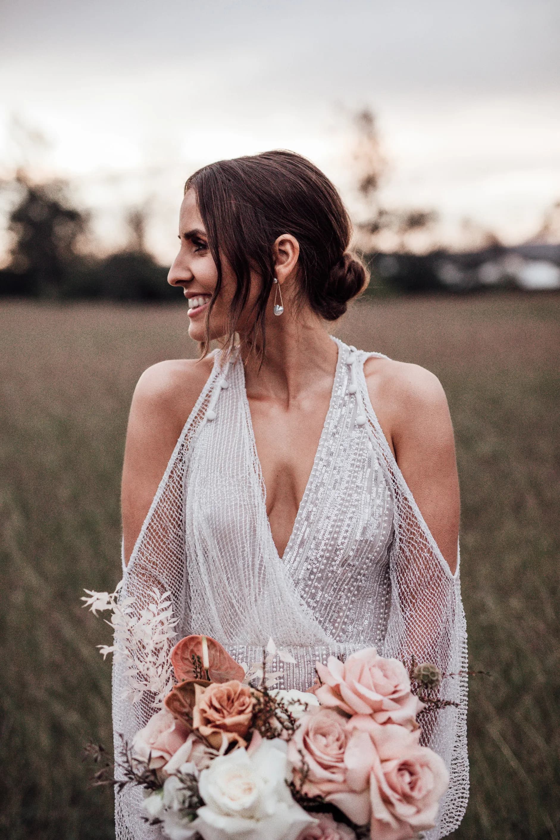 A smiling woman with dark hair in an elegant updo wearing a beaded, sleeveless white gown and drop earrings holds a bouquet of pink and white roses. She stands in a grassy field with a soft, blurred background of trees and a sky at dusk.
