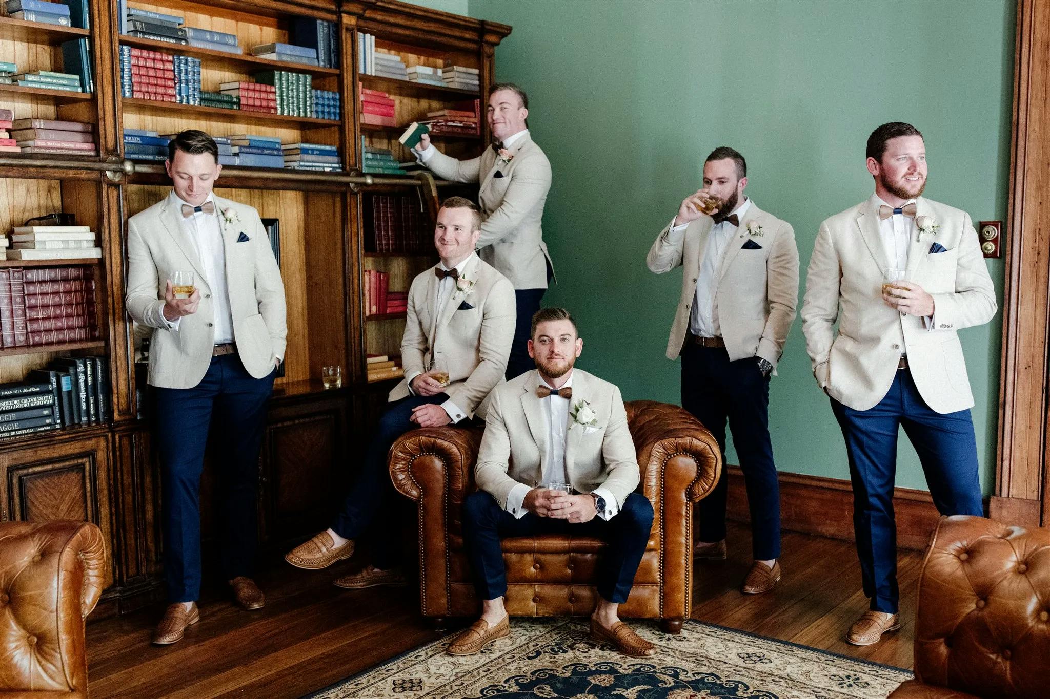 Six groomsmen in beige jackets and dark trousers pose in a vintage-style library with wooden shelves and books. Some are holding drinks, and two are sitting on a leather armchair and its armrest, creating a relaxed yet elegant atmosphere.