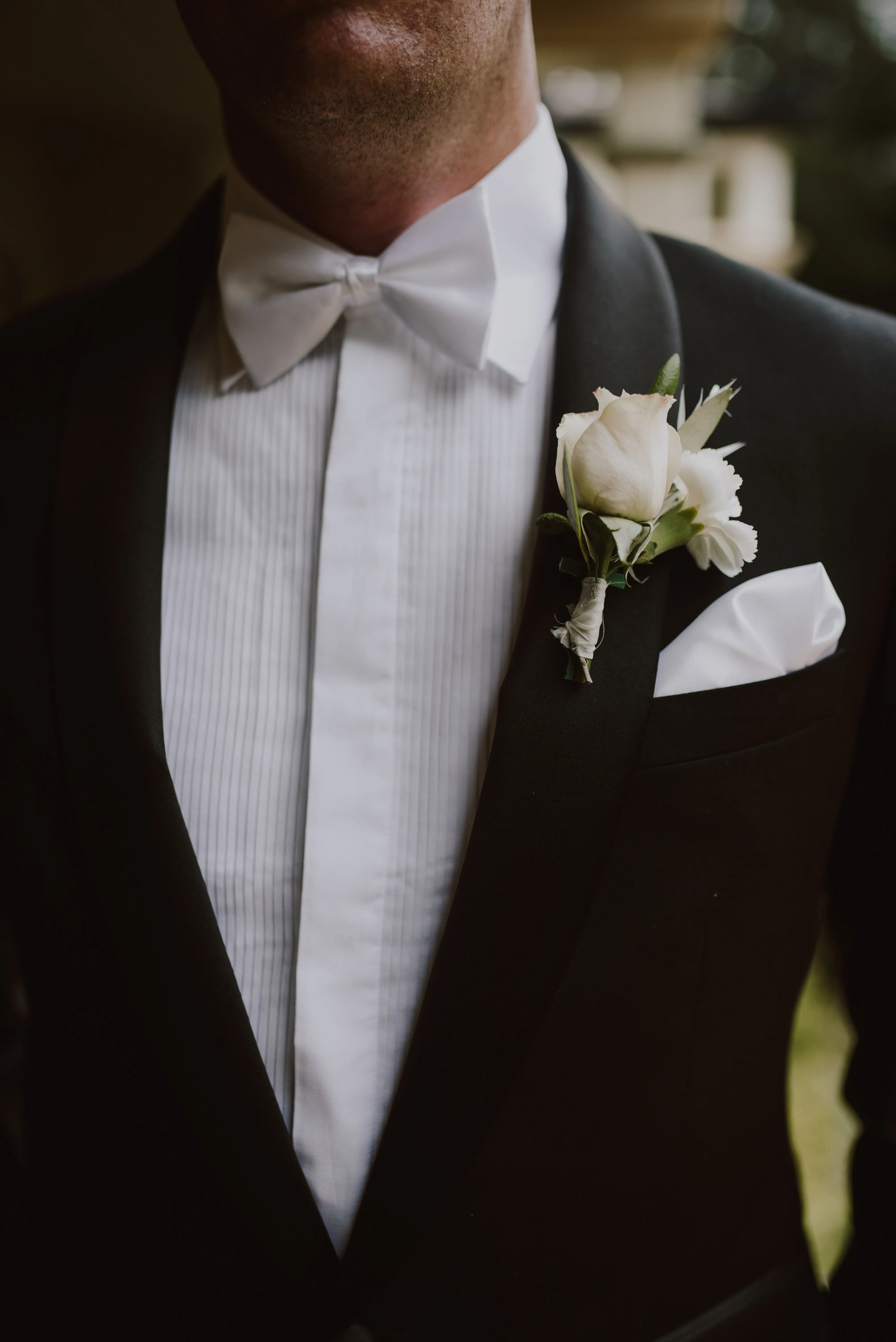 A person wearing a formal black tuxedo with a white dress shirt underneath. They have a white pocket square and a white boutonniere pinned to their lapel. The image focuses on the chest and torso, and the person is not fully visible.