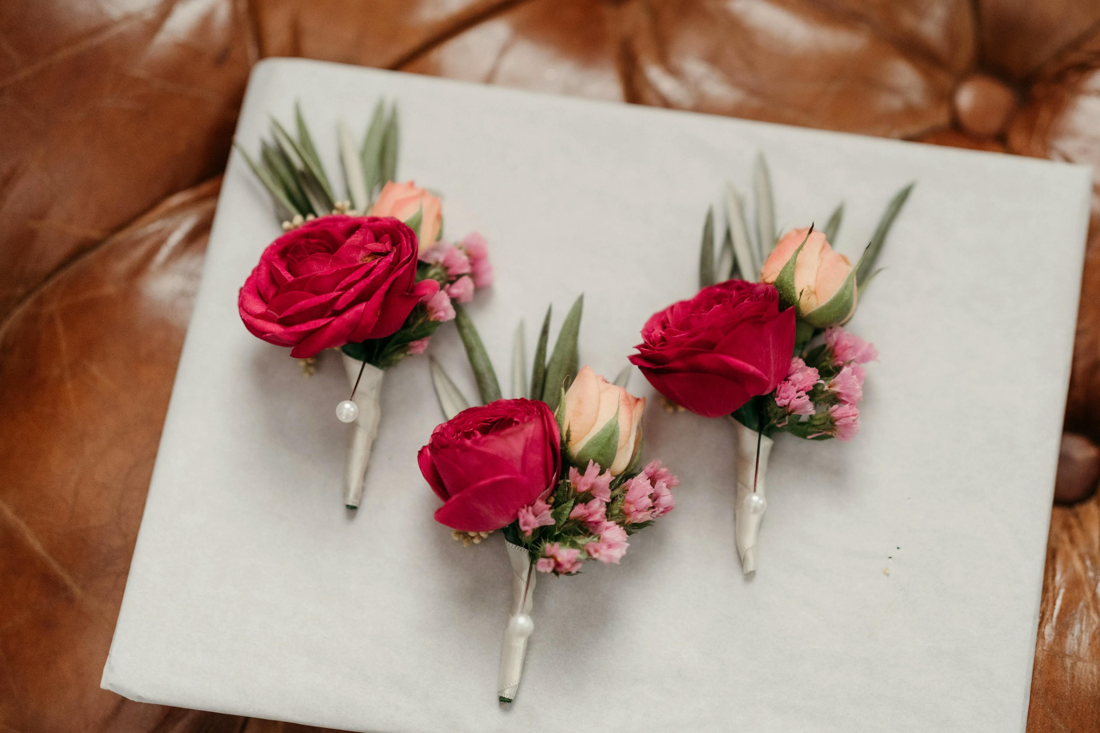 Three boutonnières arranged on a white surface, each featuring one red rose, one peach rosebud, small pink flowers, and green leaves, secured with white pins. The background is a brown leather texture.