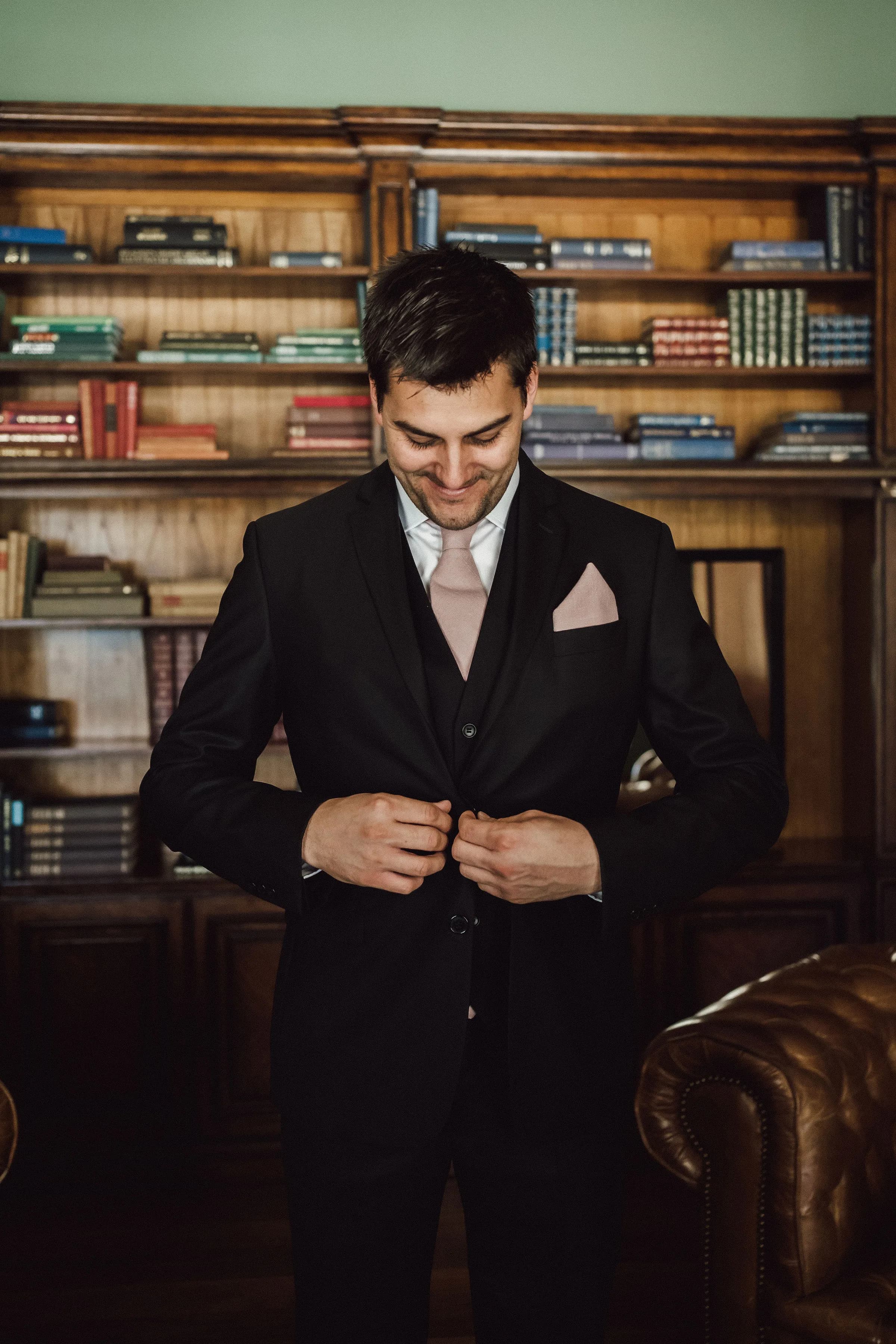 A smiling man stands in front of a bookshelf, wearing a black suit with a light pink tie and pocket square, buttoning his jacket. The background includes wooden shelves filled with colorful books and a leather armchair.