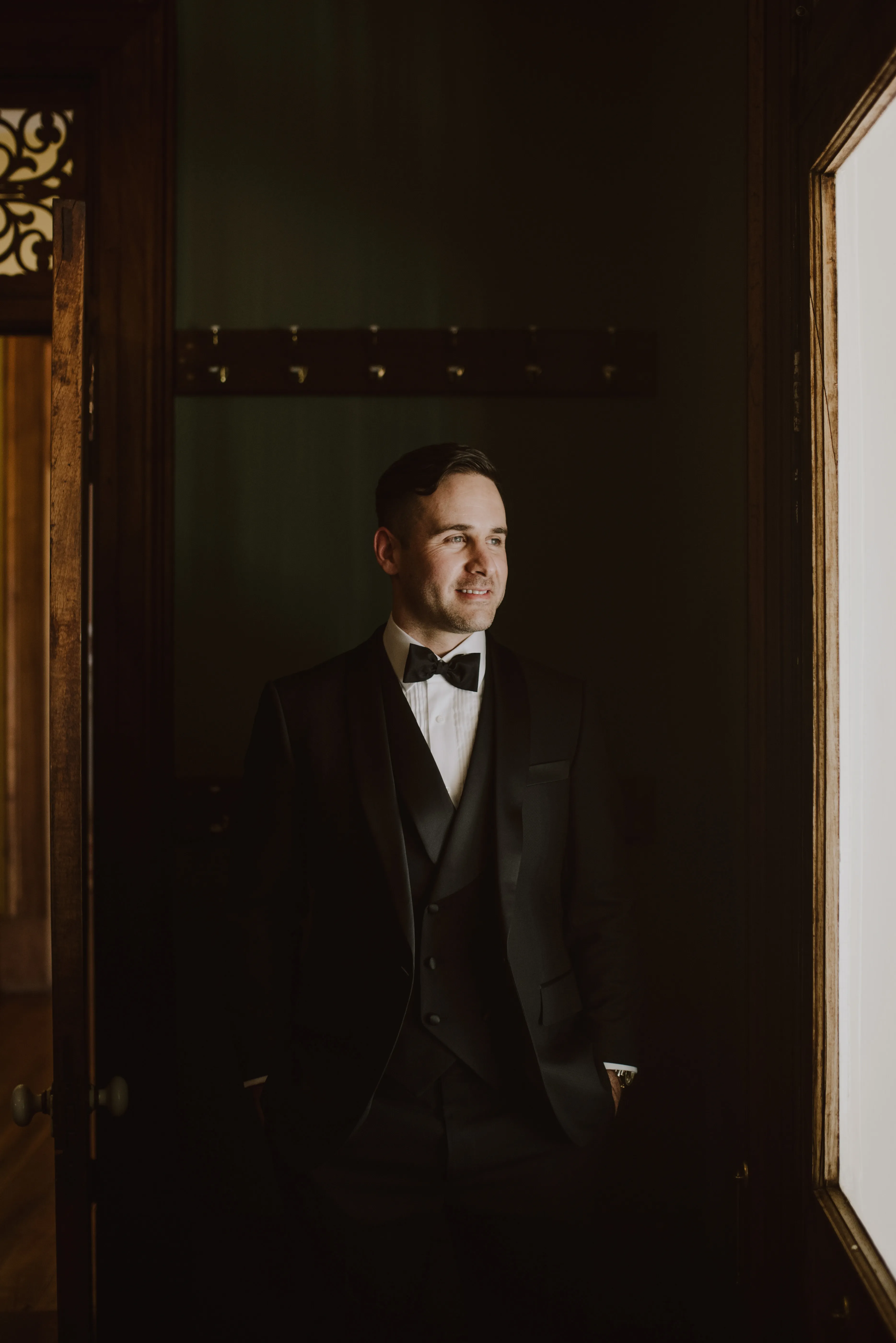 A man dressed in a dark suit and bow tie stands in a dimly lit room. Light from a window illuminates part of his face as he looks outside with a slight smile. The atmosphere is elegant and formal, with wooden doors and coat hooks in the background.