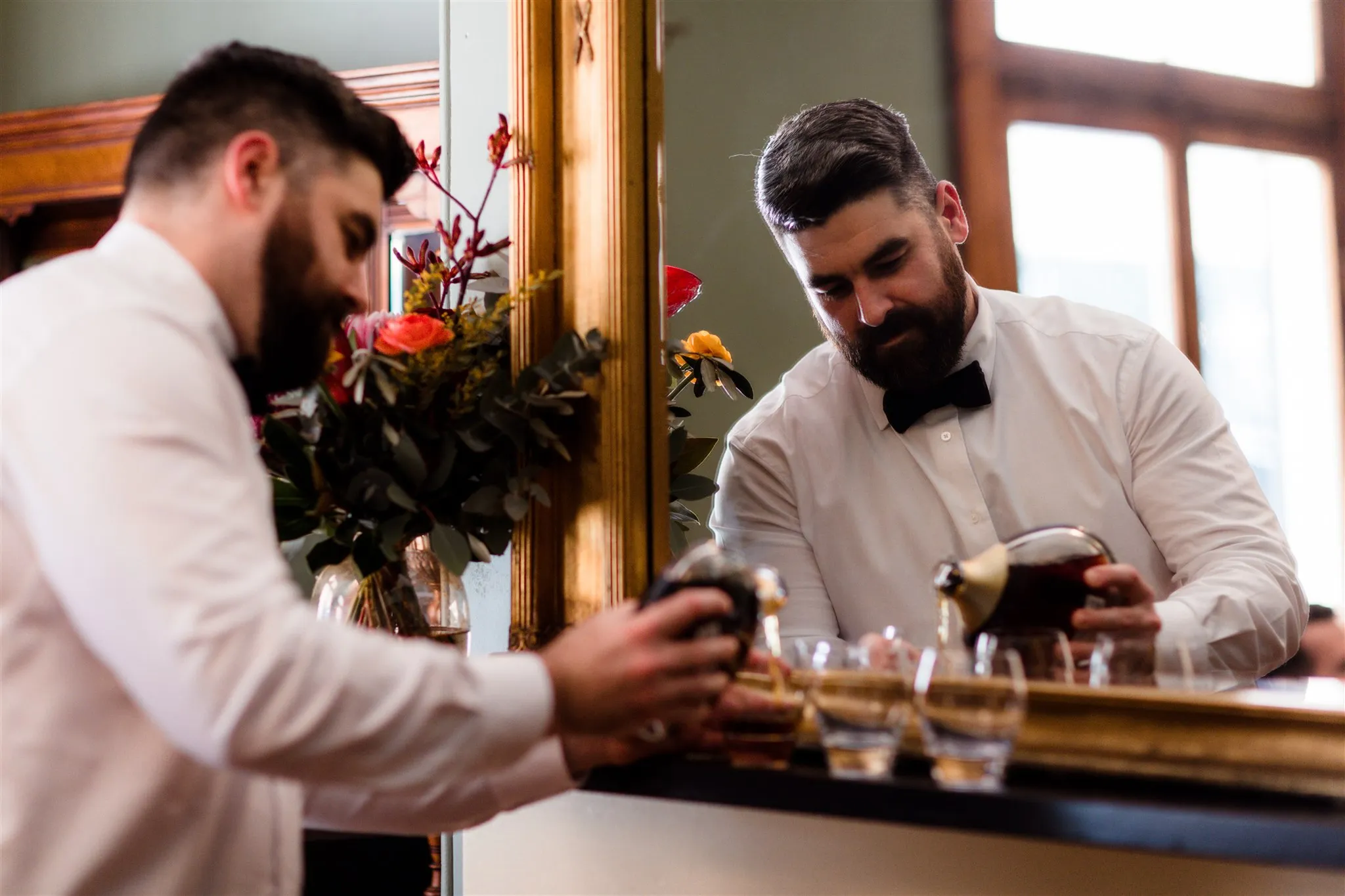 A man with a beard, dressed in a white shirt and black bow tie, is seen pouring a drink into two glasses. He is standing in front of a large mirror with a floral arrangement visible in the reflection. The setting appears to be indoors, possibly at an event.