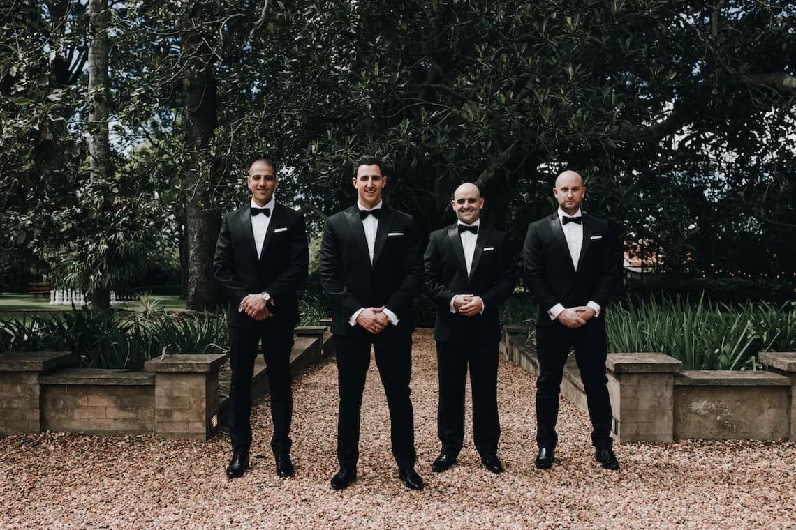 Four men in black tuxedos and bow ties stand in a gravel path outdoors, surrounded by greenery. They are evenly spaced and looking at the camera with serious expressions. Trees and plants are visible in the background.