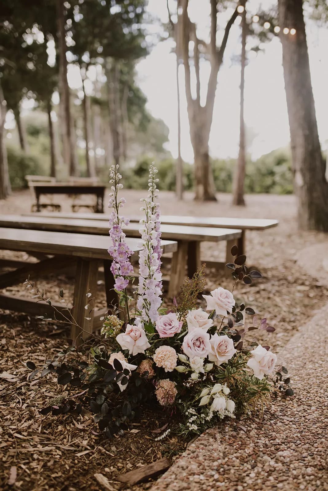 A wooden picnic table is set in a tranquil forest setting. In the foreground, there is a floral arrangement featuring pink roses, white and purple flowers, and green foliage. The ground is covered with fallen leaves and the path has small pebbles.