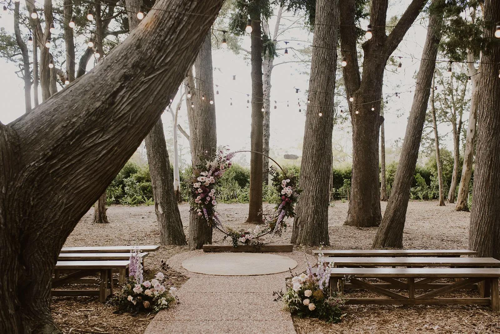 A rustic outdoor wedding ceremony setting among tall trees, featuring a circular floral arch adorned with pink and purple flowers. Wooden benches line both sides of the aisle, which is decorated with additional floral arrangements. String lights hang above.